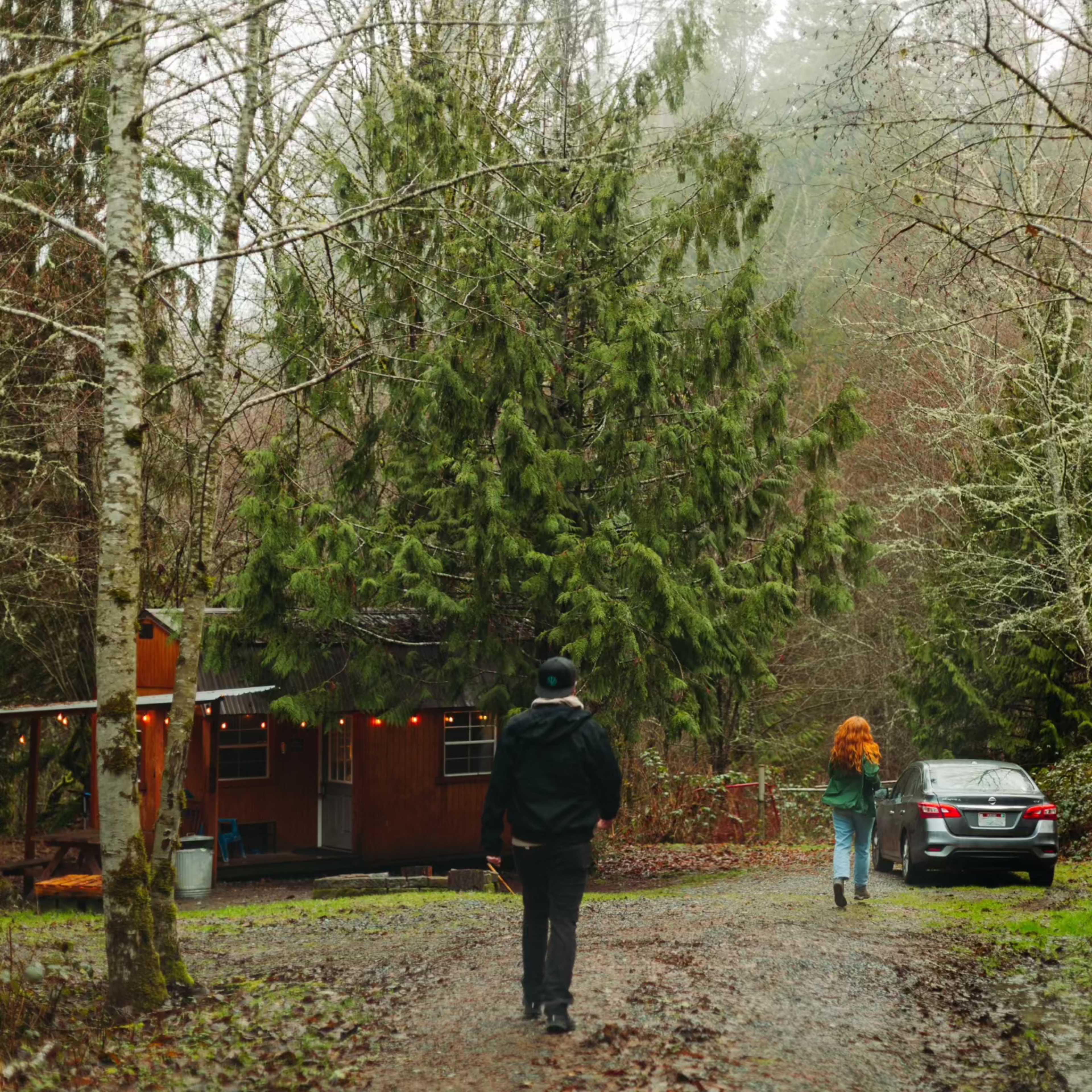 Two individuals walk along a gravel path toward a cabin surrounded by tall trees in a misty forest setting.