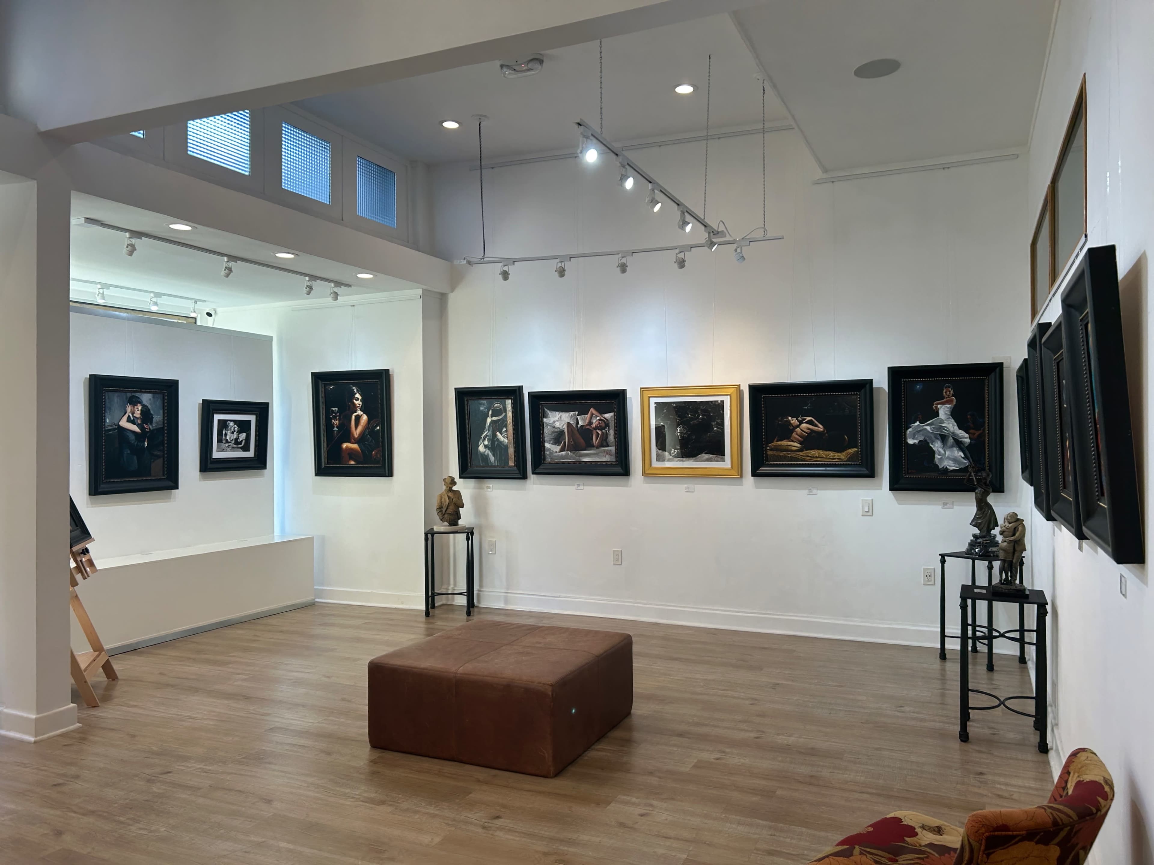 The image shows an art gallery with framed photographs displayed on white walls, a brown ottoman in the center, and a small sculpture on a stand to the left.