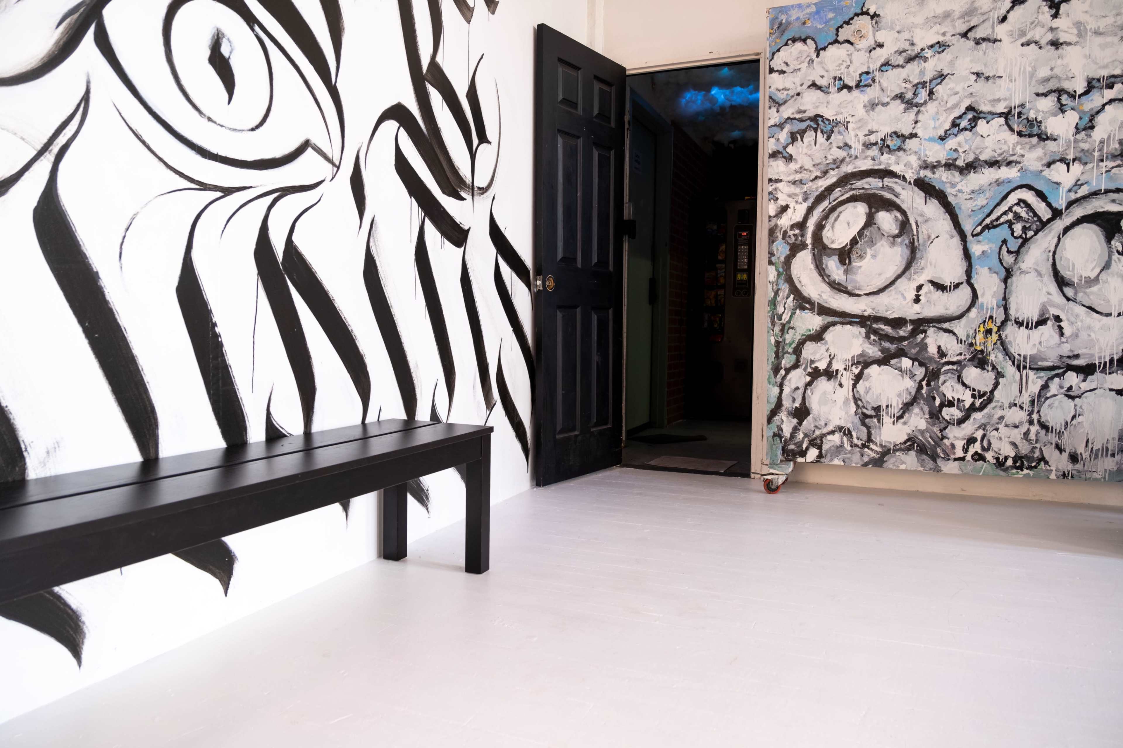 A minimalist gallery space with black and white abstract murals on the walls, a wooden bench in the foreground, and an open doorway leading to another room.