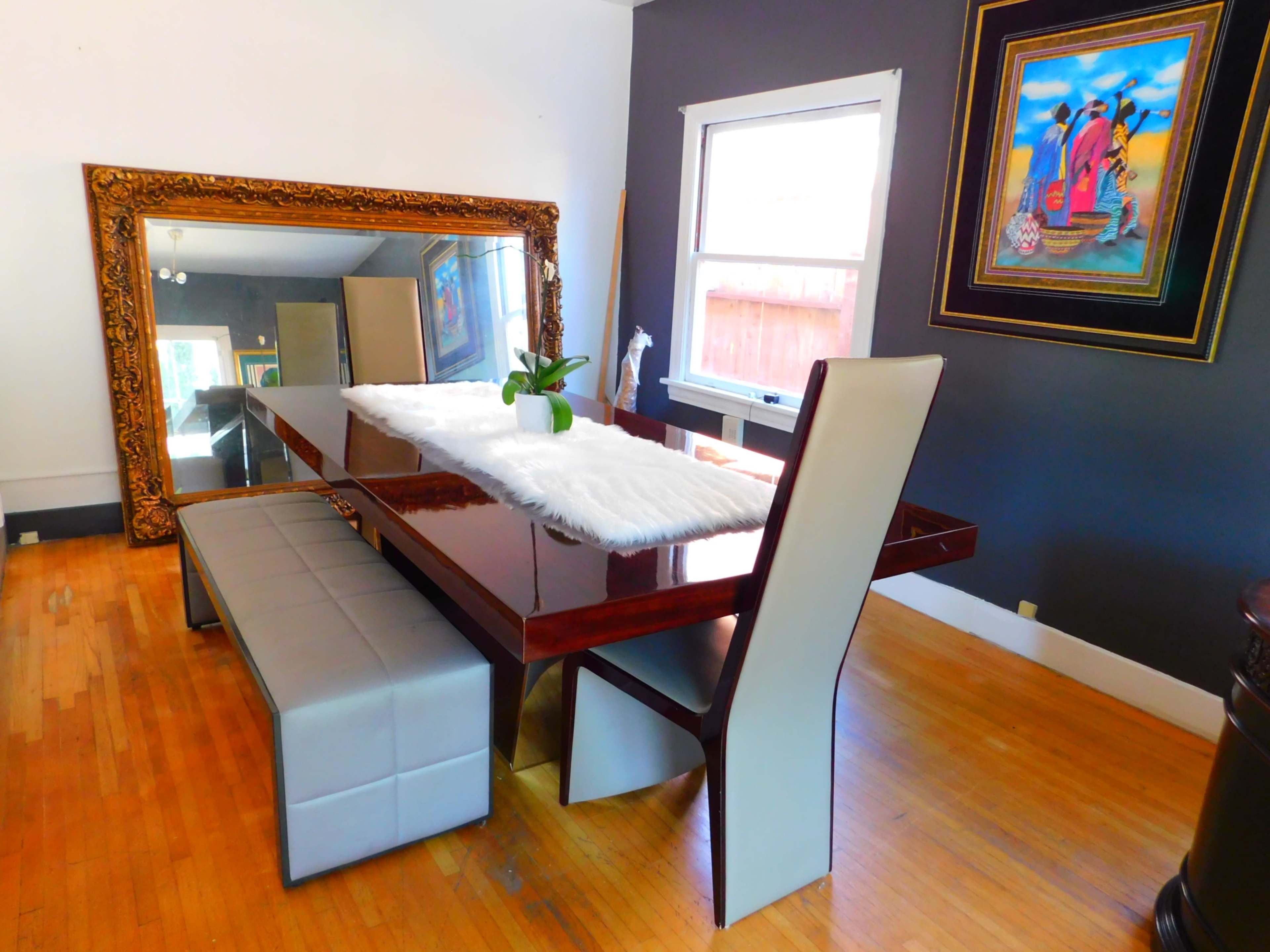 A wooden dining table with a white runner and a potted plant is accompanied by a gray bench and surrounded by walls painted dark gray, with a large ornate mirror and colorful artwork in the background.