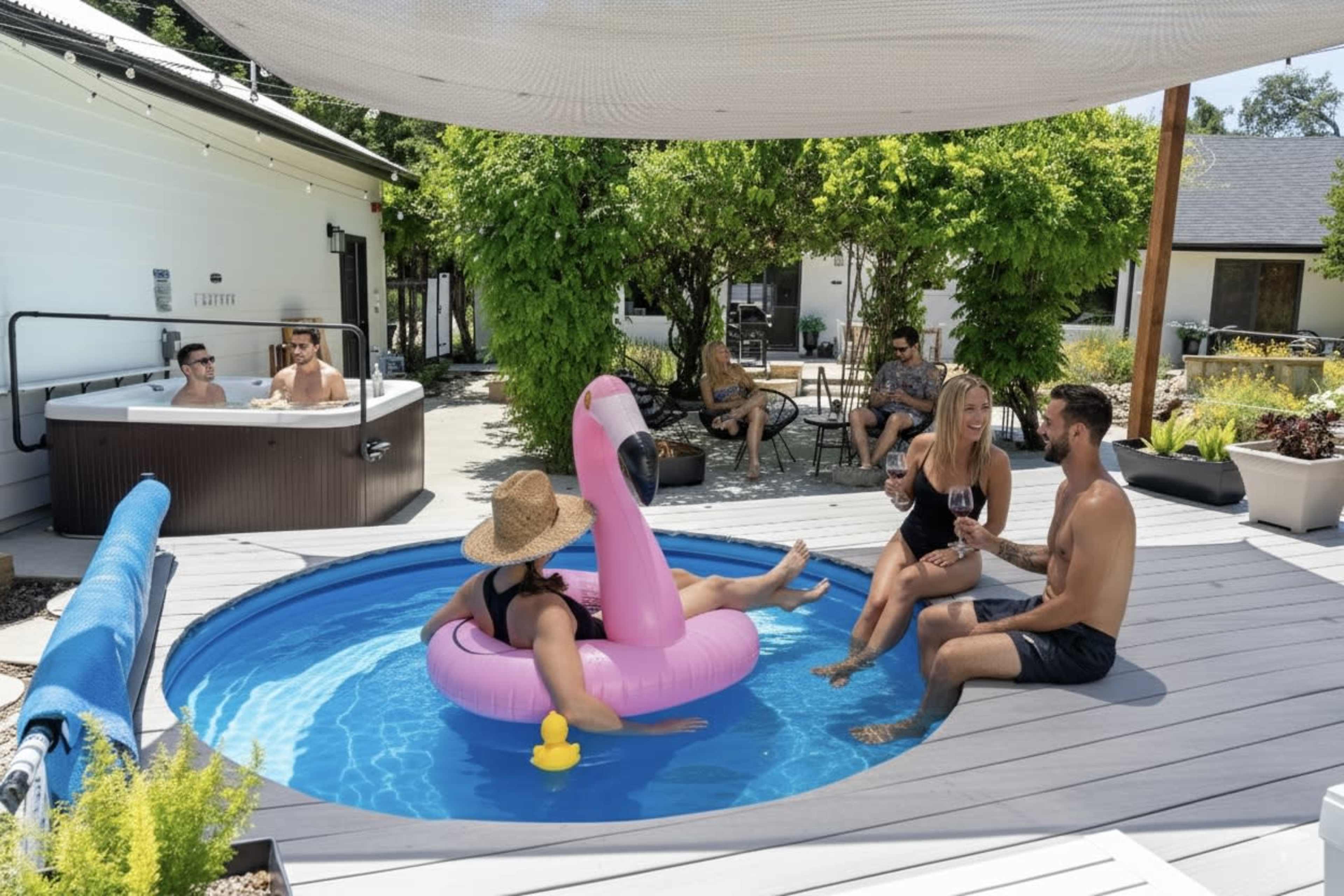 A group of five people relaxes by a pool, with one person on a pink flamingo float, while two others enjoy drinks and a hot tub is visible in the background.