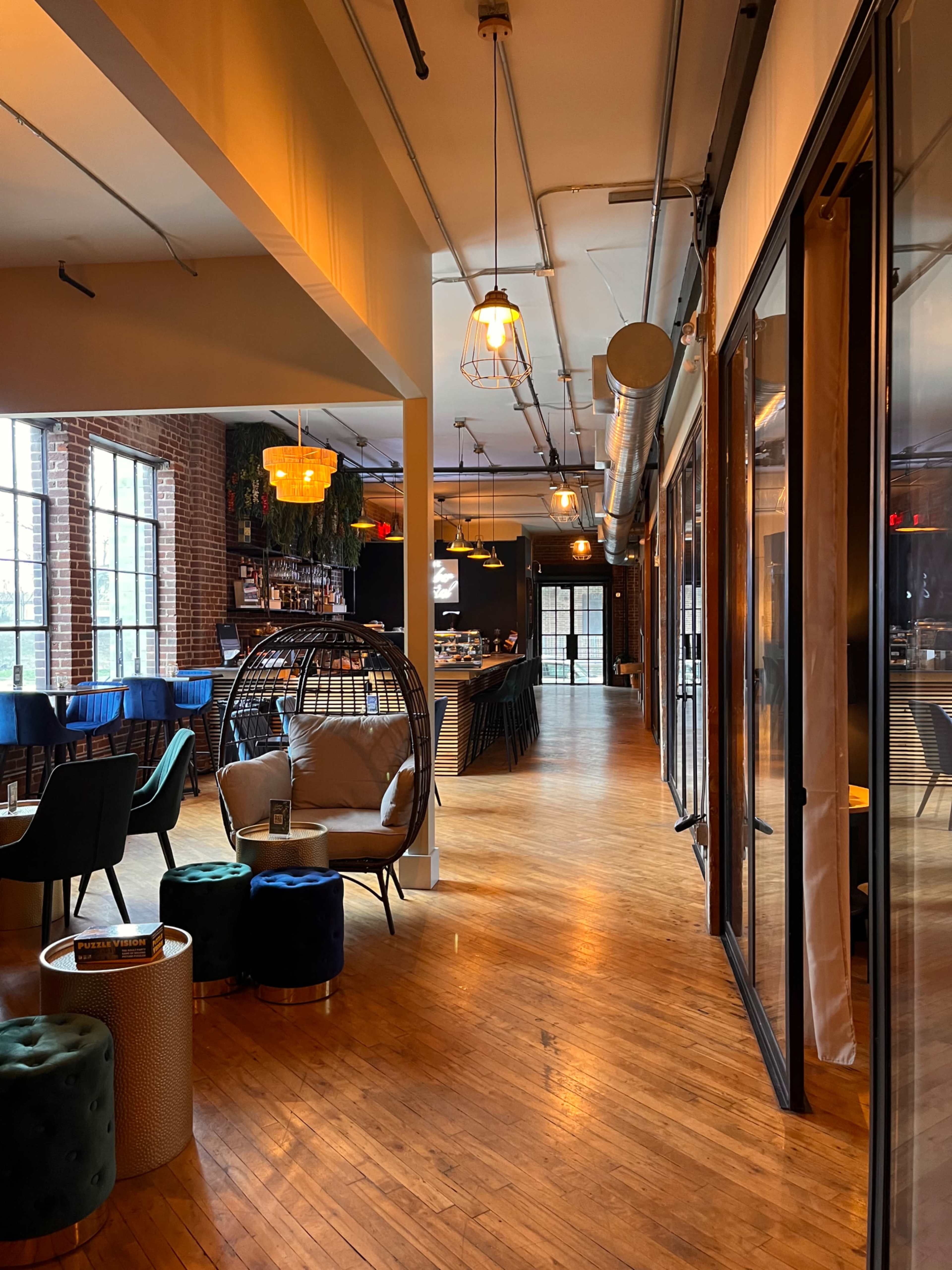 A stylish interior of a modern café or lounge with wooden flooring, industrial-style lighting, and a seating area featuring a hanging chair and colorful stools.