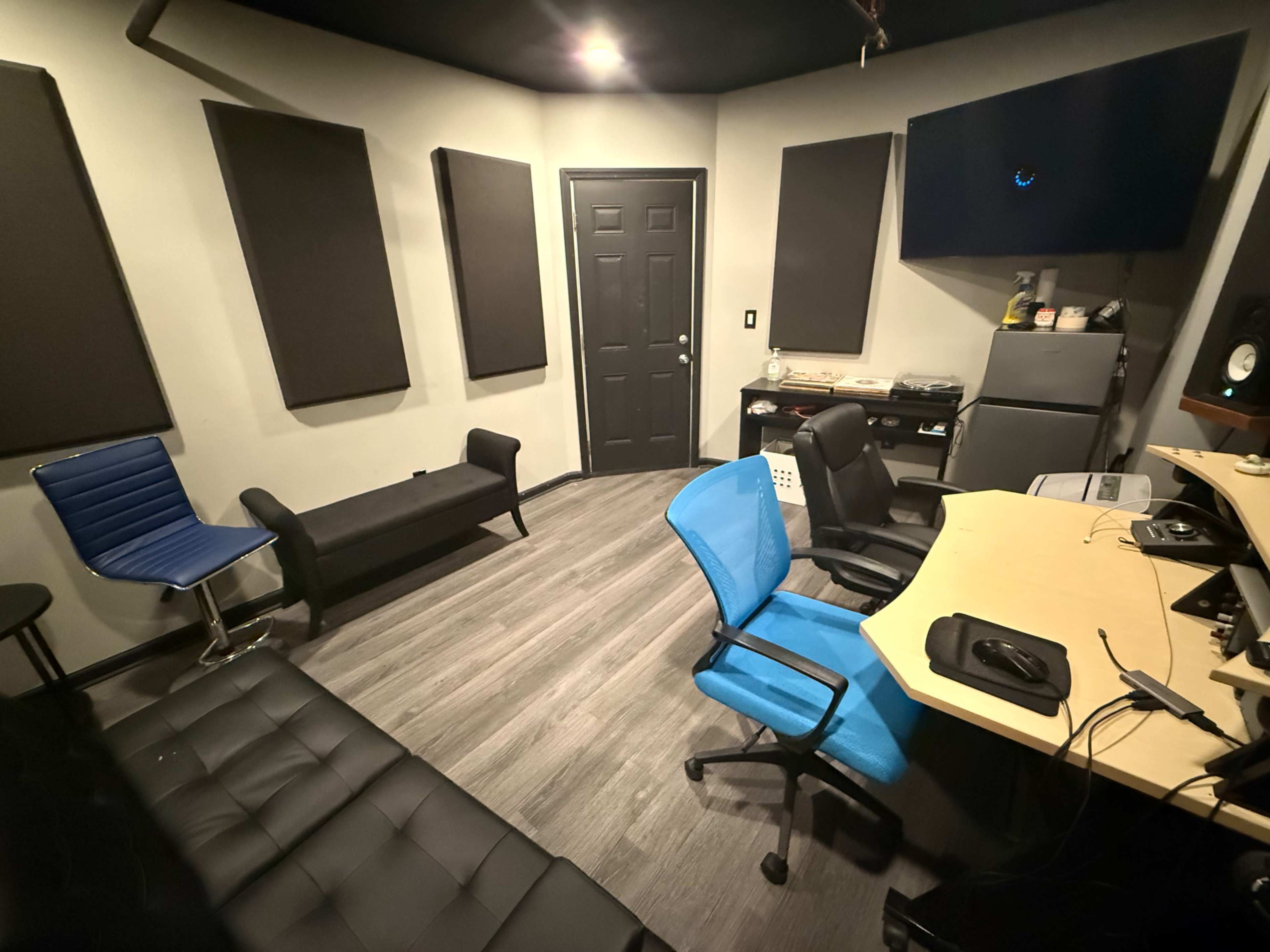 The image shows a recording studio equipped with soundproofing panels, a desk with a computer setup, and various seating options, including two chairs and a couch.