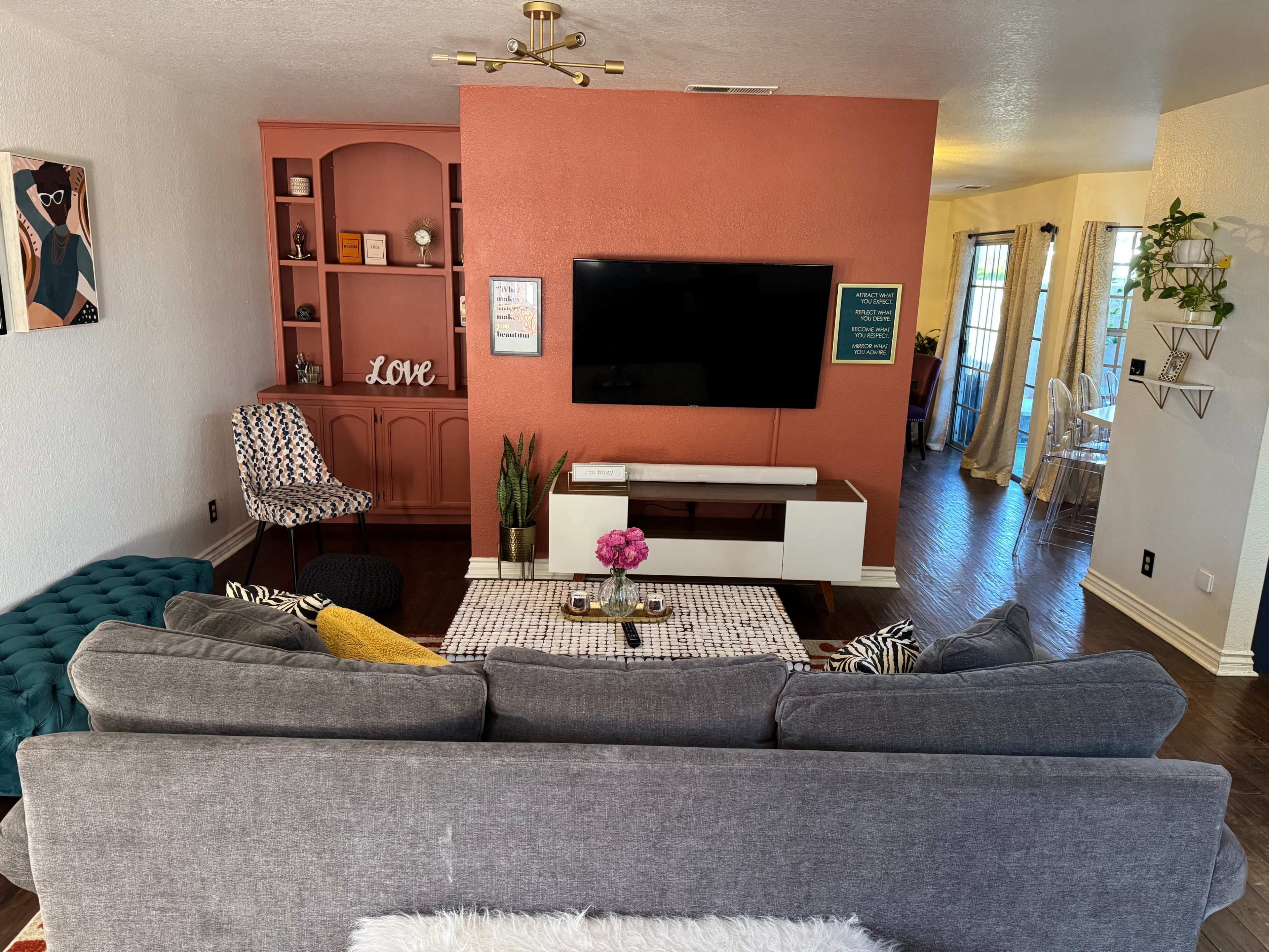 The image shows a living room featuring a gray sofa, a white coffee table with a pink flower centerpiece, and a mounted television on a coral accent wall adjacent to a bookshelf and a chair.
