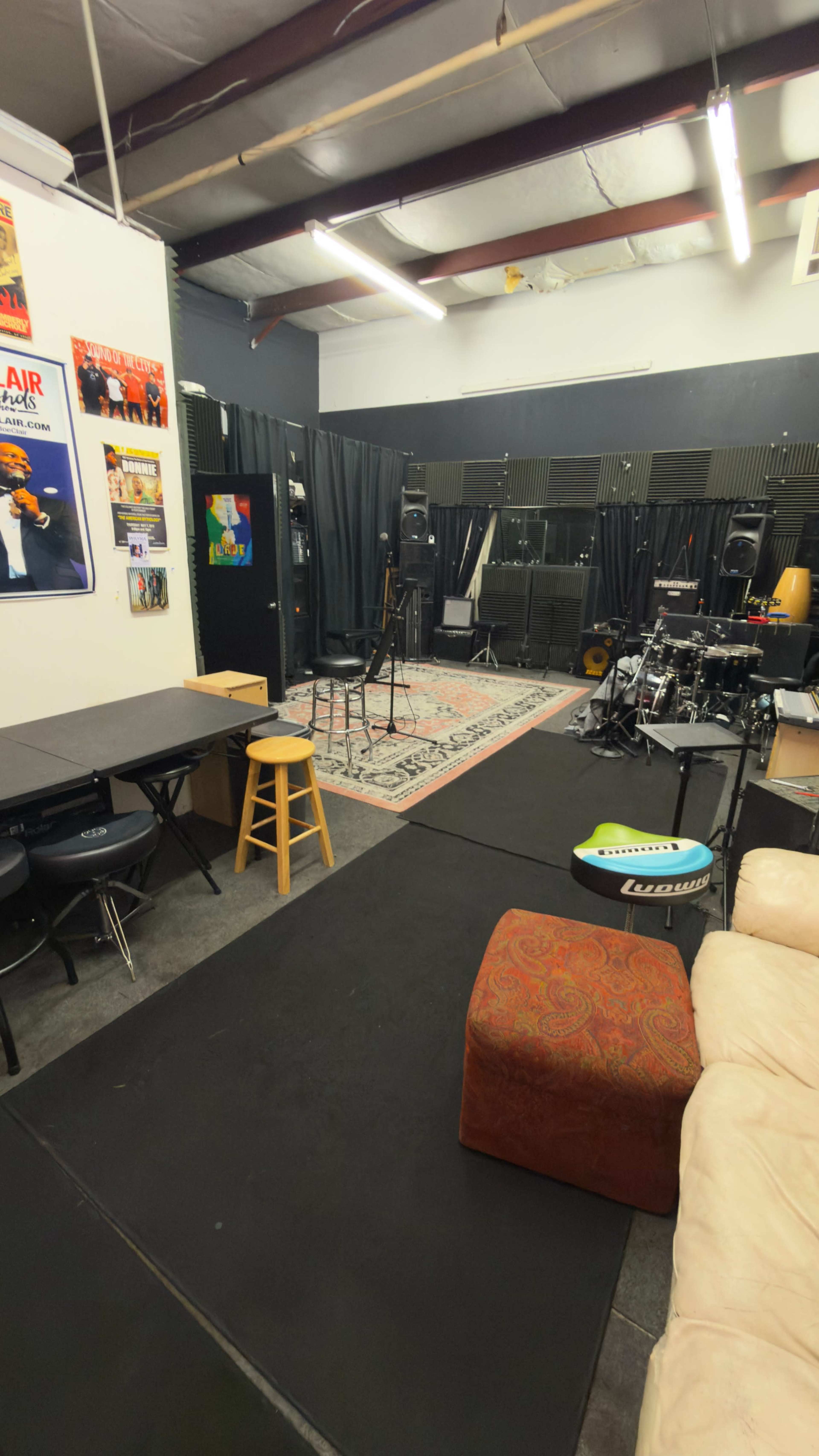 The image shows a music rehearsal space featuring black curtains, musical instruments, a patterned rug, and a lounge area with seating.
