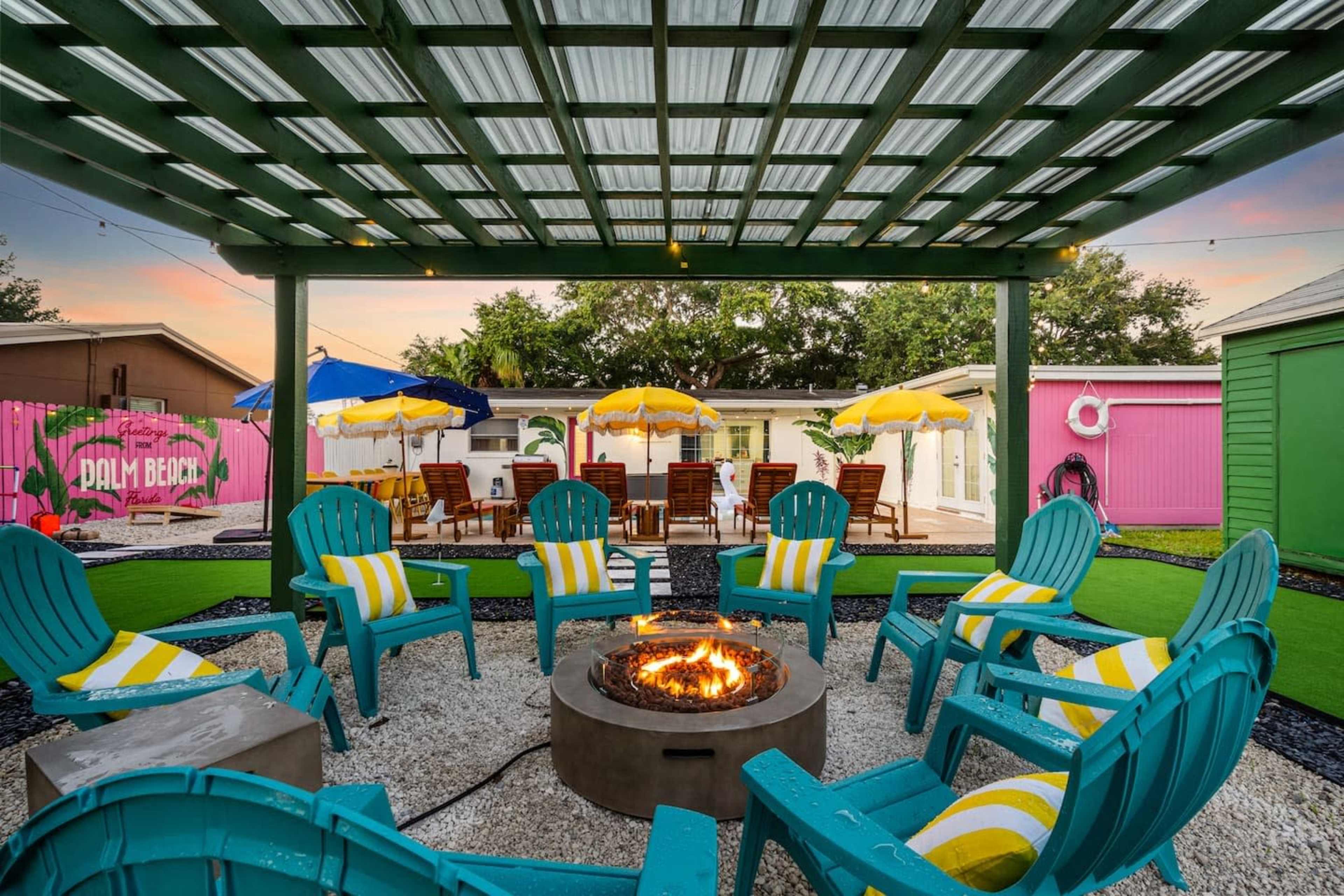The image shows a vibrant outdoor seating area with teal Adirondack chairs arranged around a fire pit under a green pergola, surrounded by colorful umbrellas and a pink building in the background.
