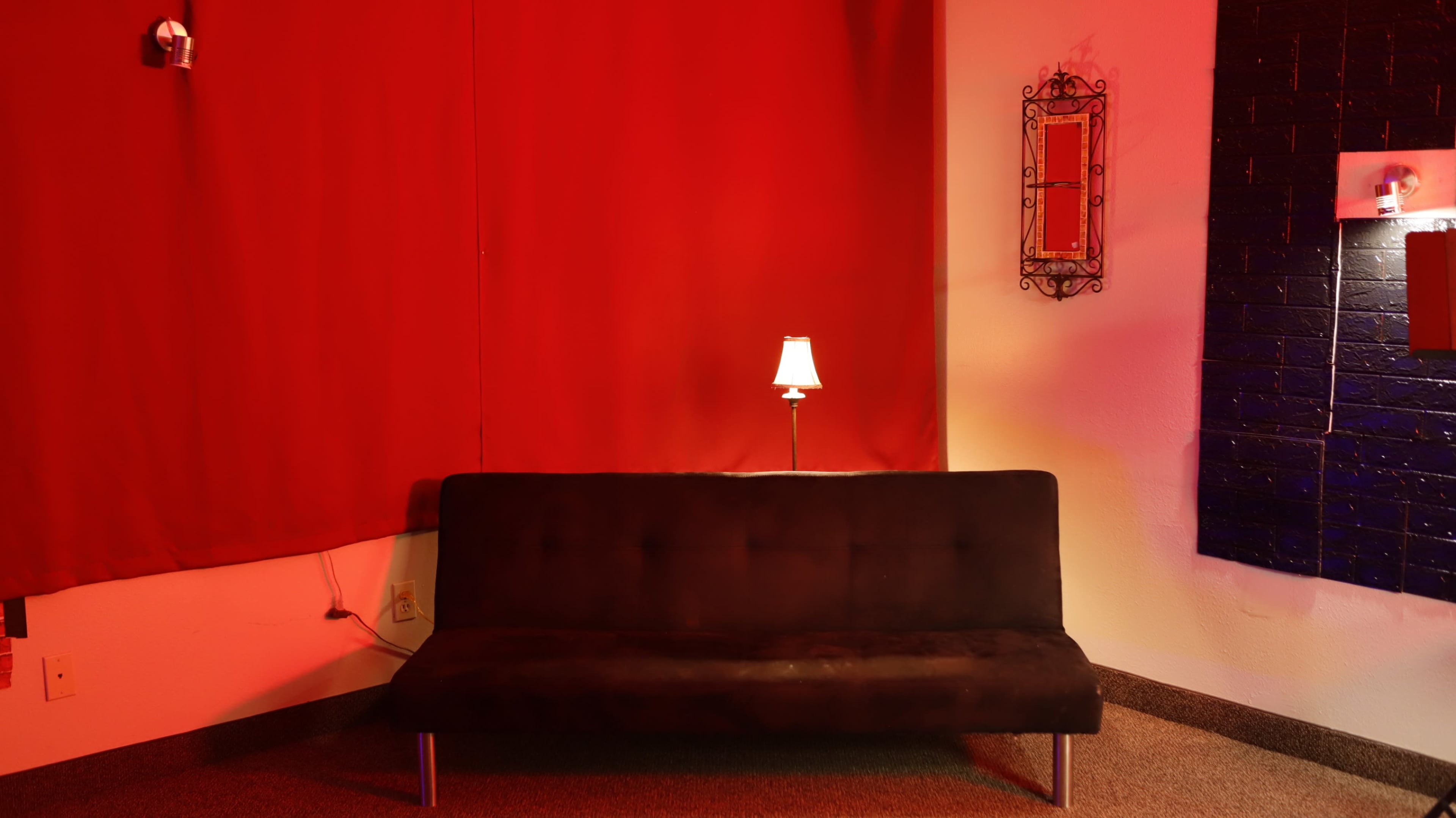 A dark sofa is positioned against a wall with red curtains and a small lamp illuminating the space.