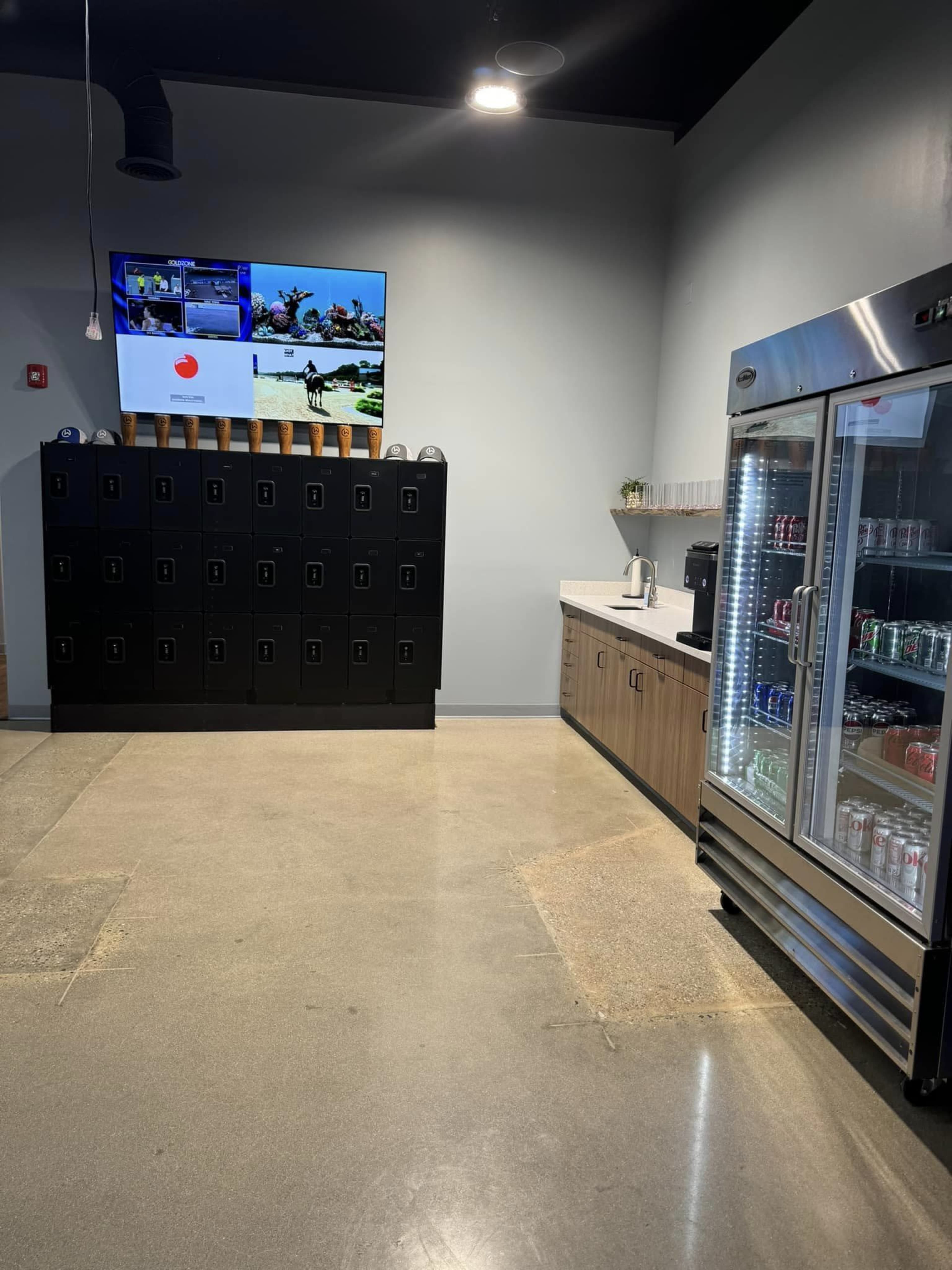 A modern break area featuring black lockers, a beverage fridge, a coffee station, and two television screens mounted on the wall.