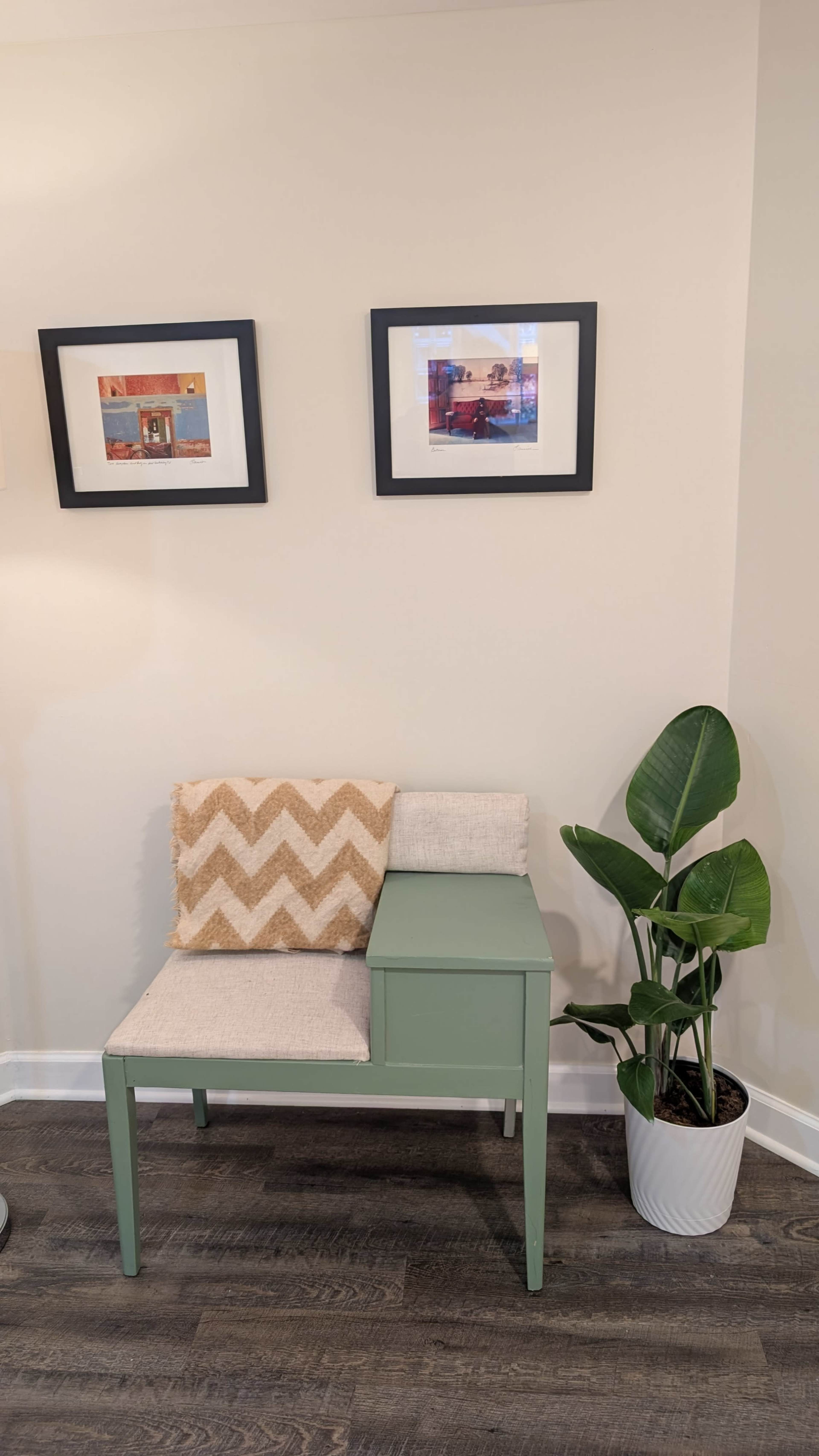 The image features a green accent bench with a light-colored cushion, placed against a wall adorned with two framed pictures, next to a leafy potted plant.