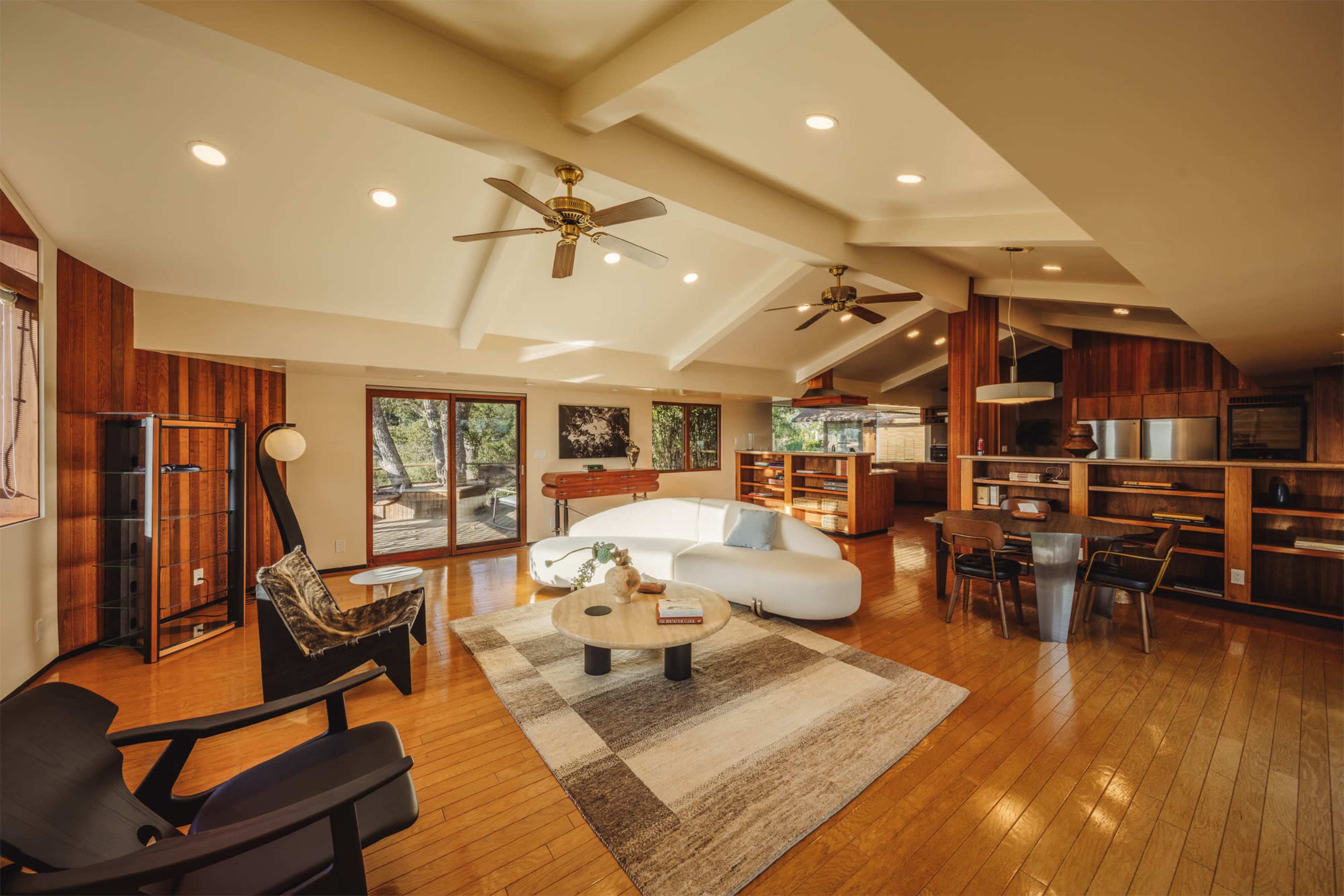 The image shows a spacious living area featuring wooden flooring, a white curved sectional sofa, two ceiling fans, and large windows that open to an outdoor deck.