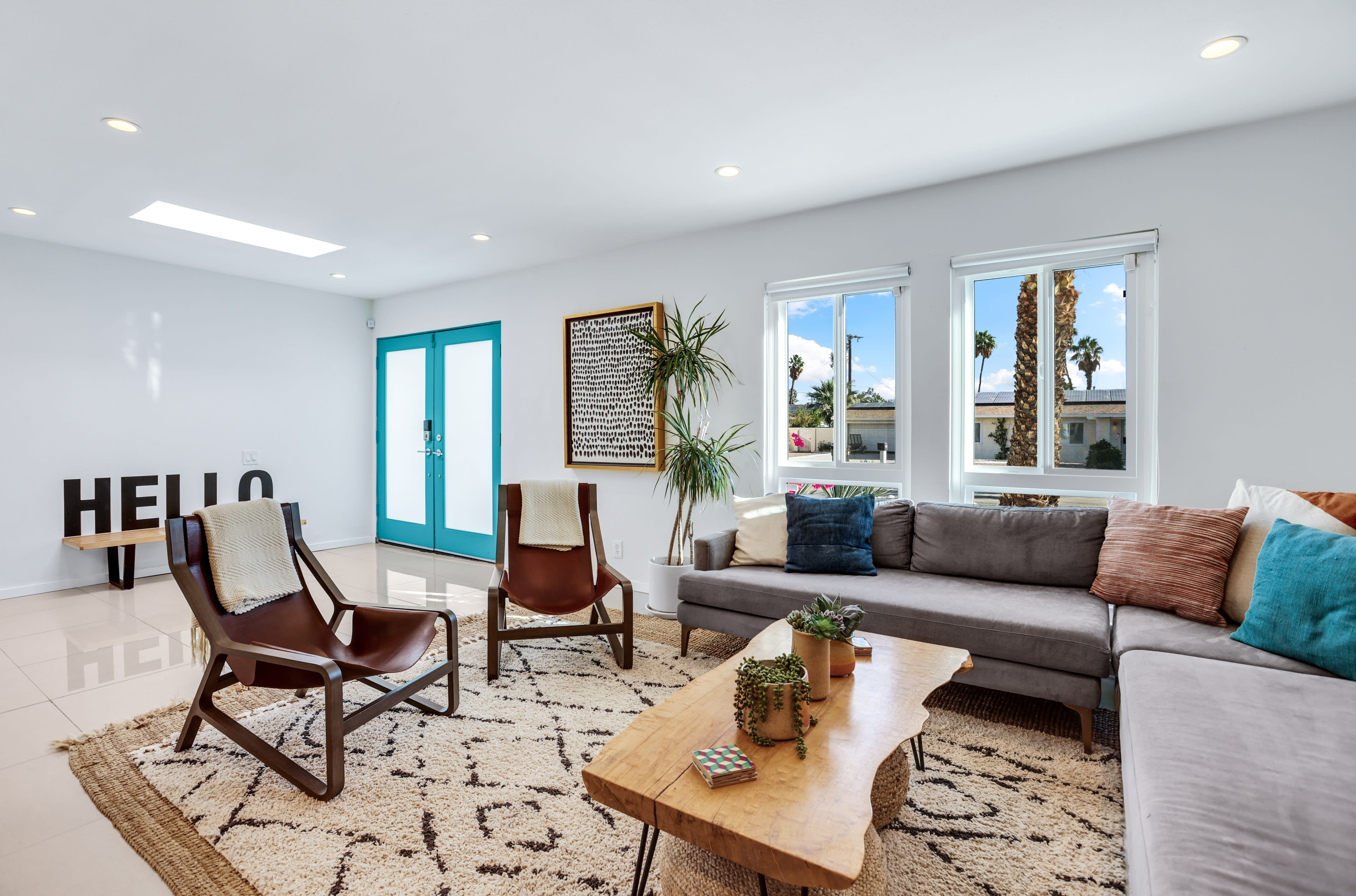 A bright living room with a gray sectional sofa, two wooden chairs, a large patterned rug, and a wooden coffee table, featuring a double door entrance with teal accents and windows that allow natural light in.