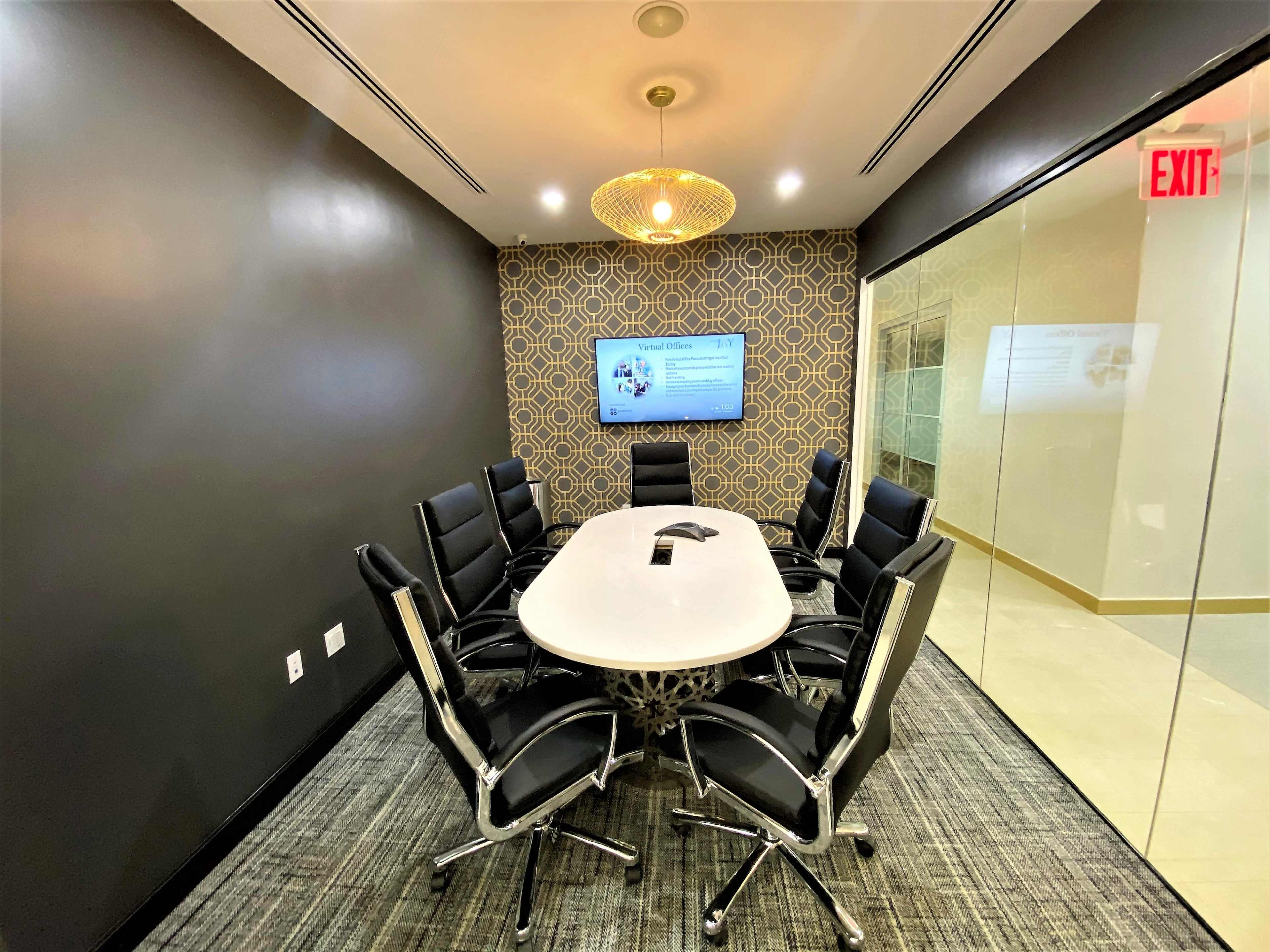 A modern conference room features a long white table surrounded by black chairs, with a decorative wall and a screen displaying information.