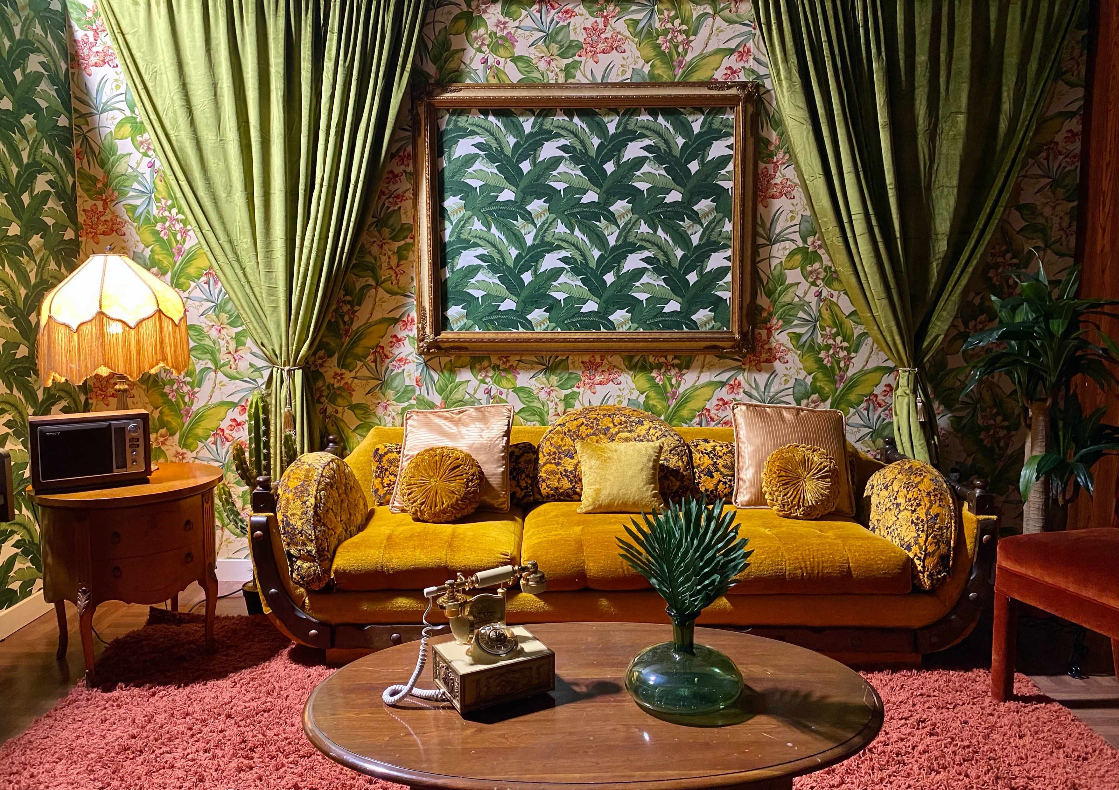 The living room features a yellow, patterned sofa with green curtains, a round table, a vintage telephone, and a decorative lamp, all set against a floral wallpaper backdrop.