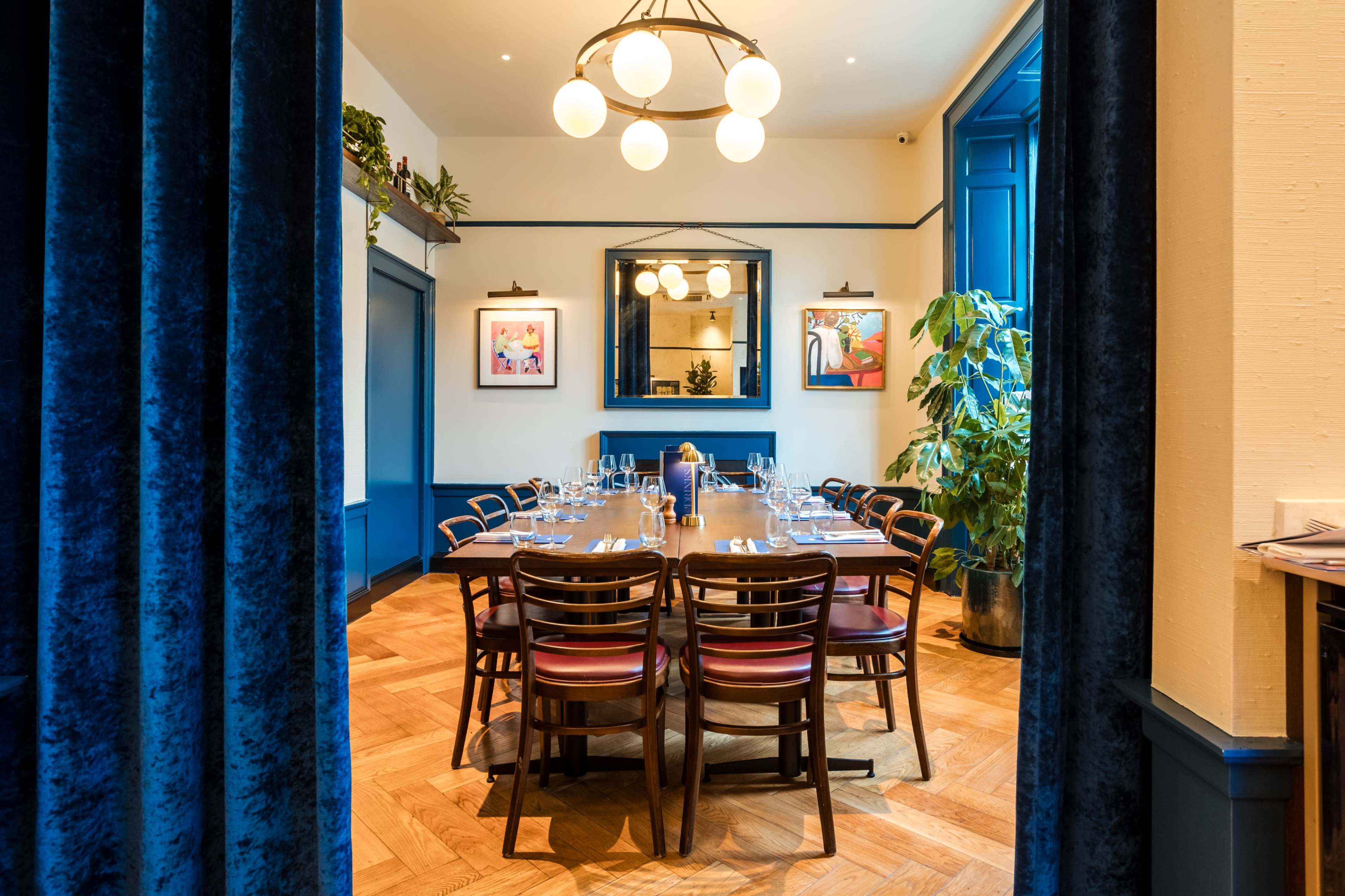 The image shows a dining room with a long wooden table set for a meal, surrounded by wooden chairs, and decorated with plants and artwork on the walls.