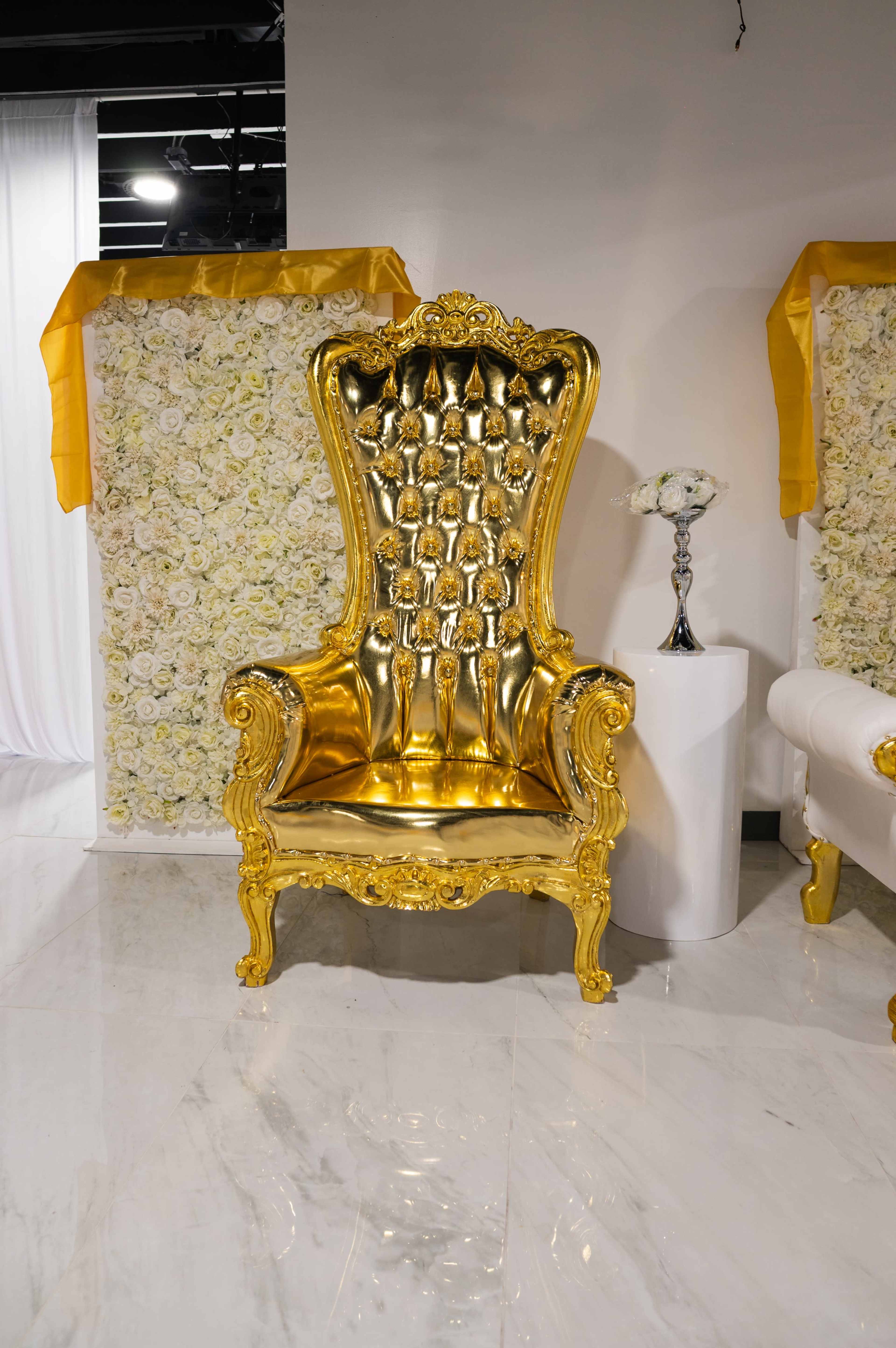 The image features an ornate golden throne with a tufted backrest, positioned against a backdrop of artificial white flowers.