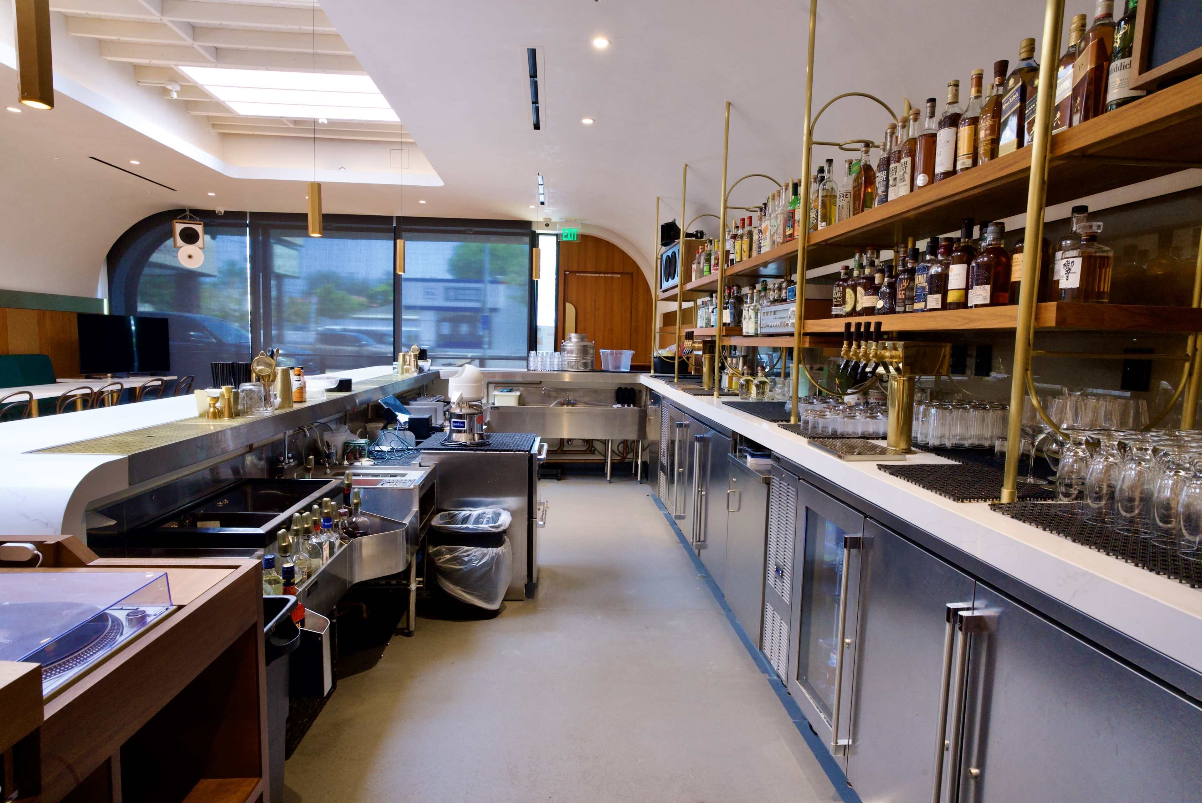 The image shows a modern bar with a polished metal counter, shelves filled with various bottles, and a clean workspace for preparing drinks.
