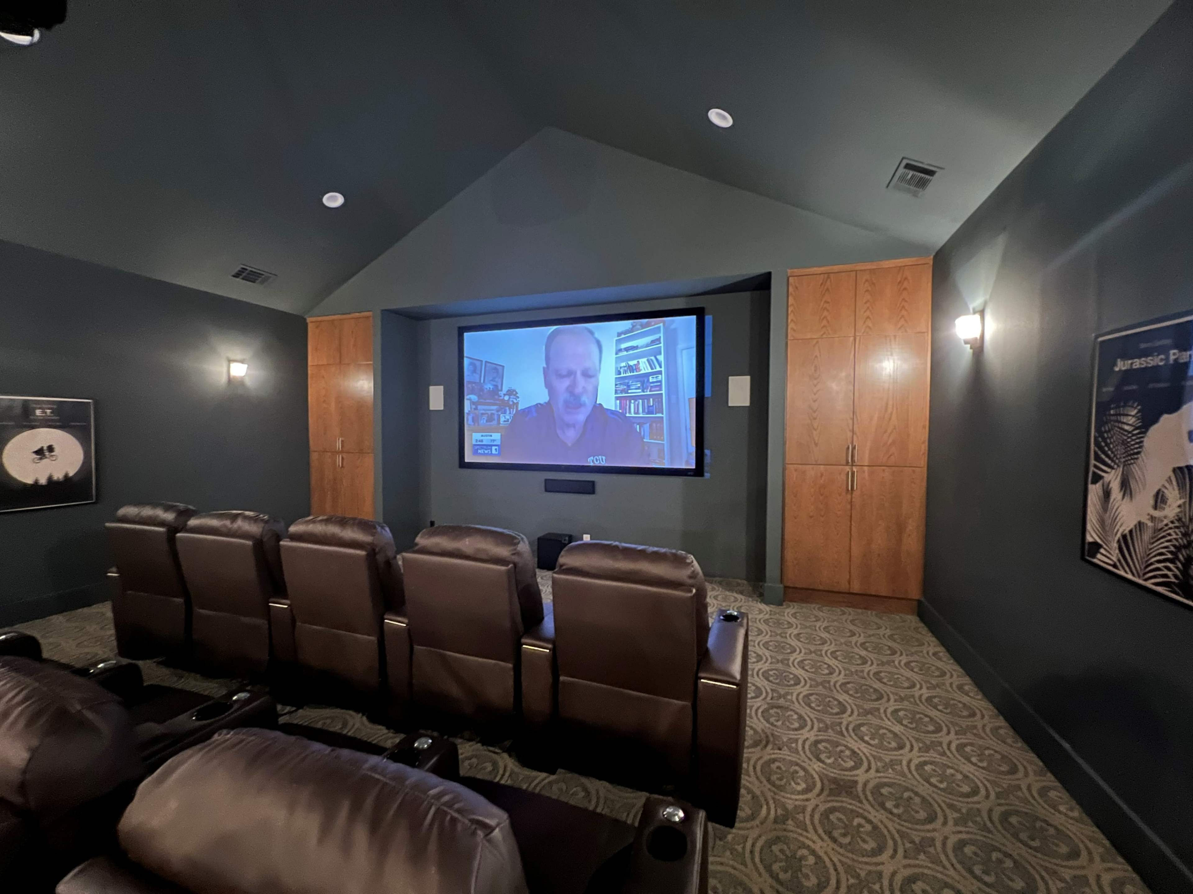 A home theater features leather recliner seats facing a large screen displaying a video call with a man in a blue shirt.