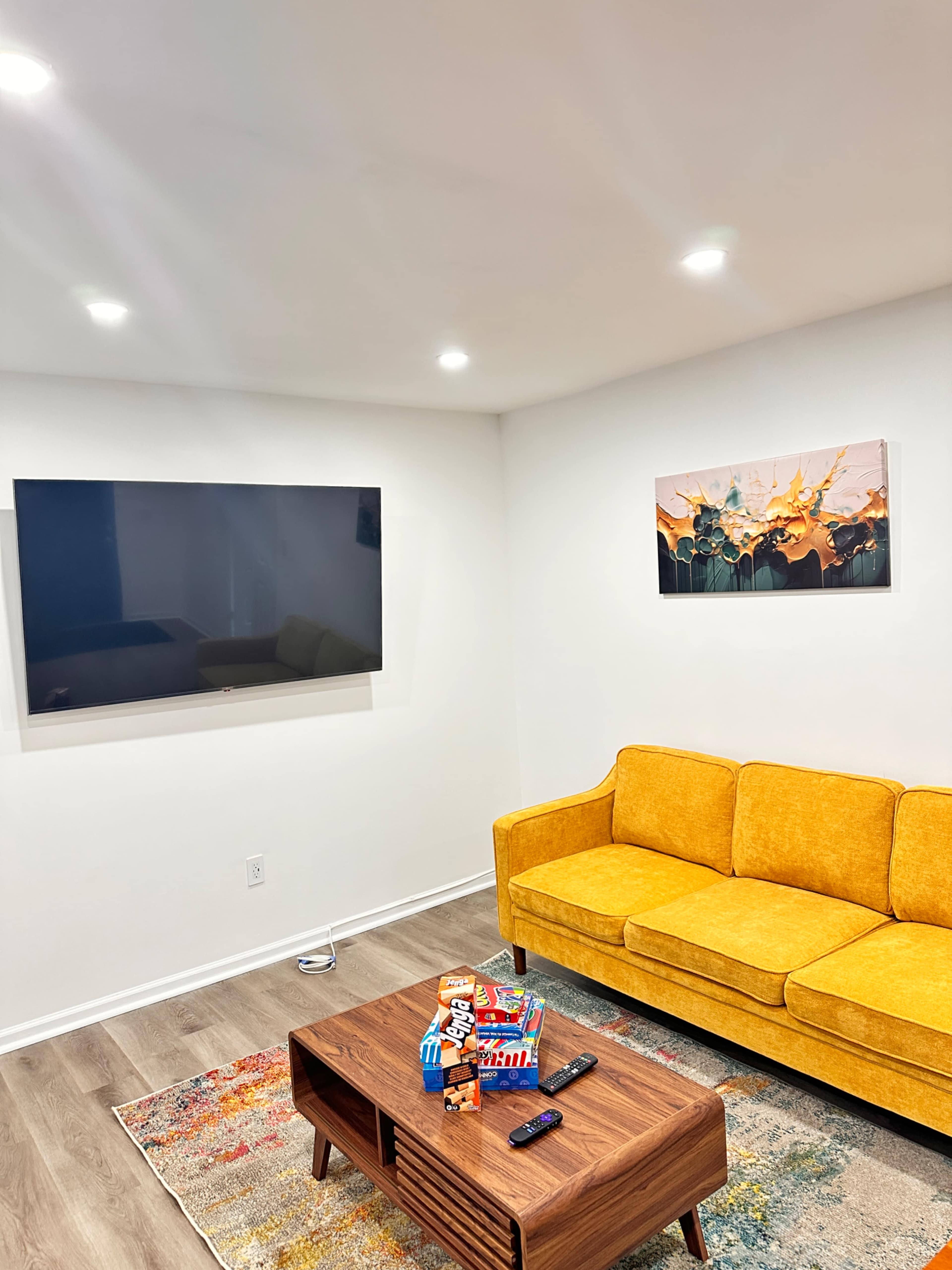 The image shows a modern living room with a yellow couch, a wooden coffee table with decorations, a mounted flat-screen TV on the wall, and a colorful area rug.