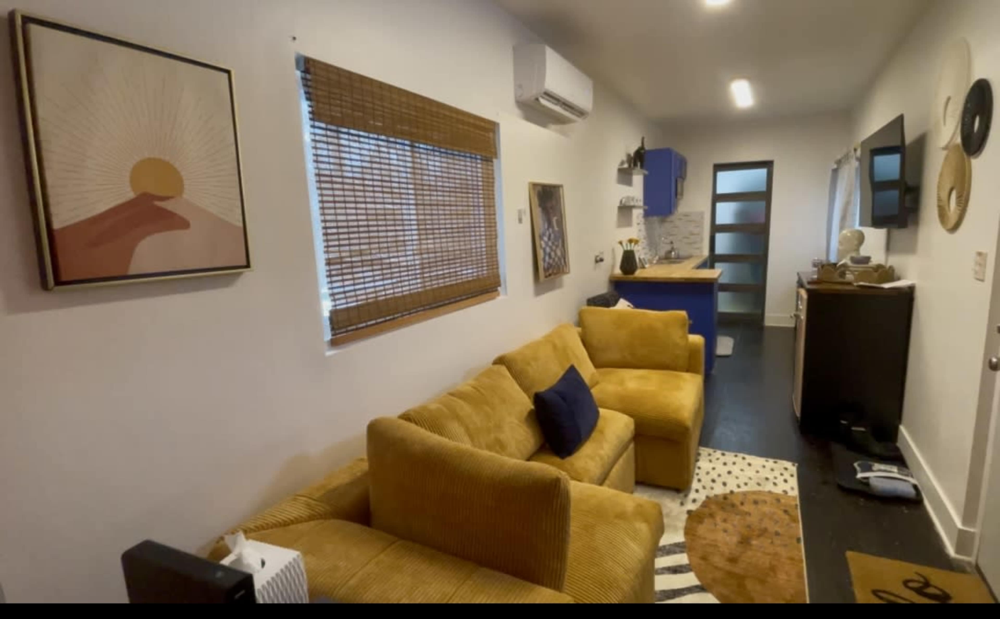 The image shows a cozy living area with a yellow sectional sofa, modern artwork on the wall, and a small kitchen area in the background.