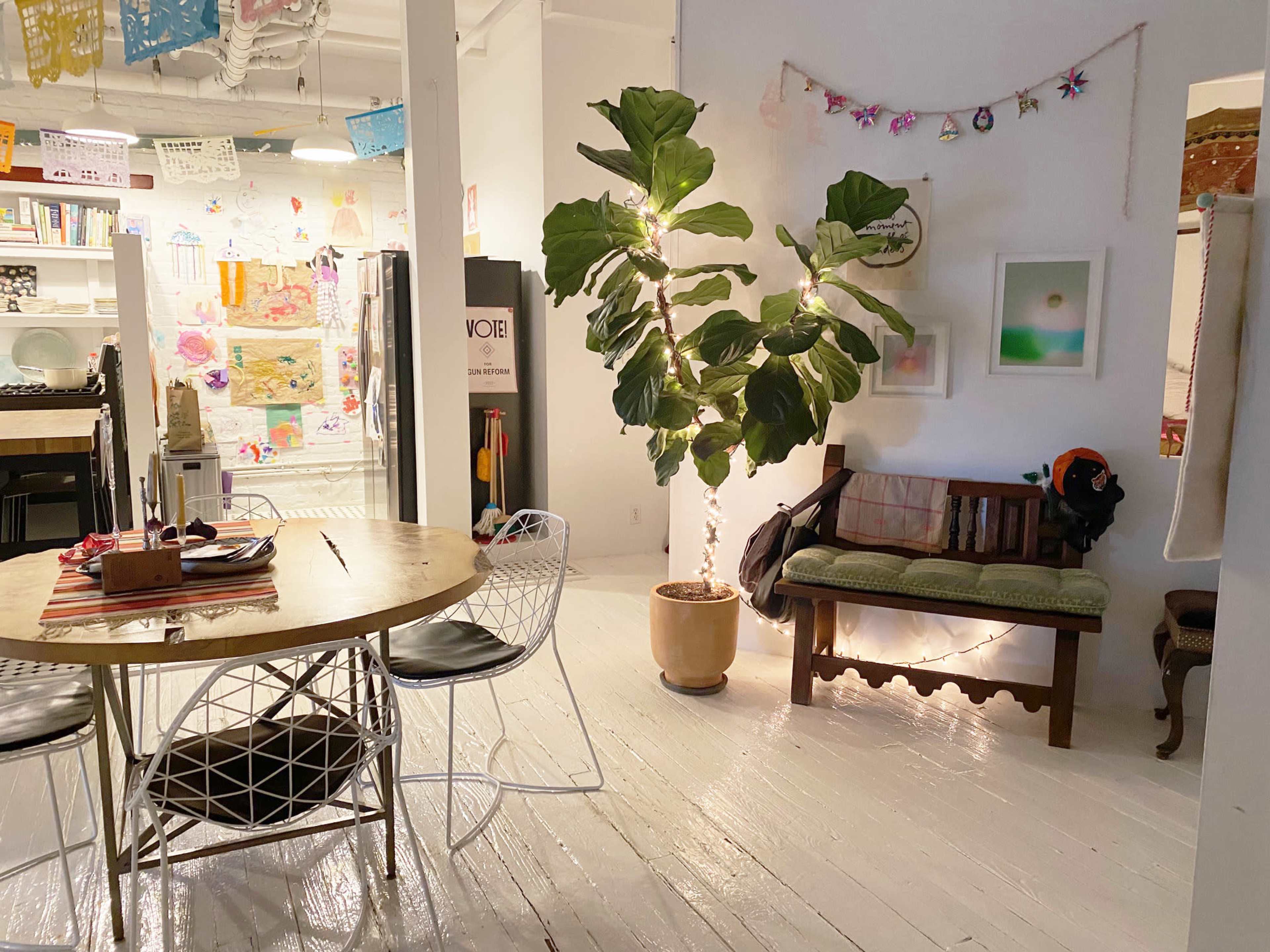 A brightly lit room with a dining table and chairs, a large houseplant in a pot, and a sofa decorated with string lights.