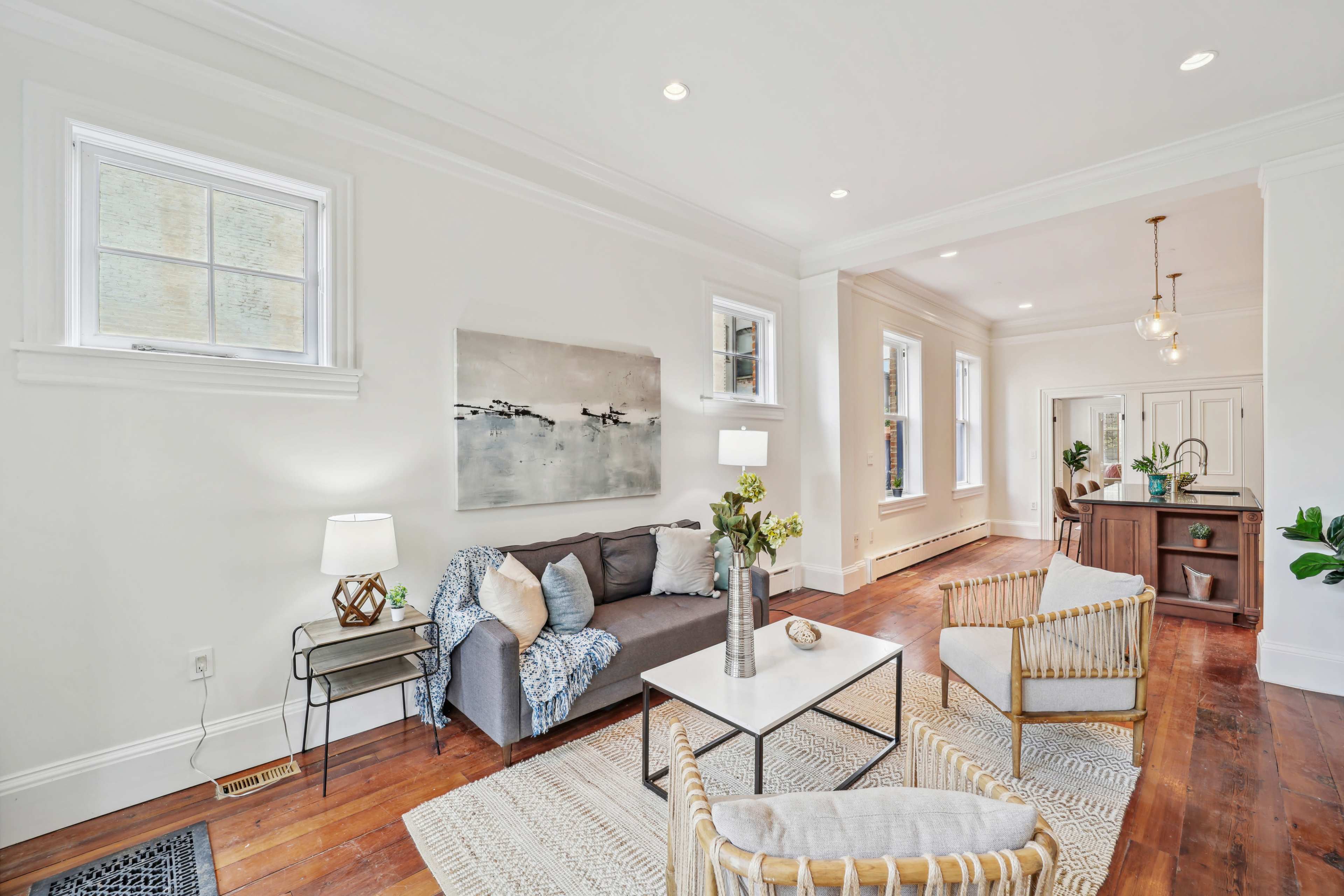 A bright, modern living room with a gray sofa, a light area rug, and a wooden coffee table, featuring large windows that allow natural light to illuminate the space.