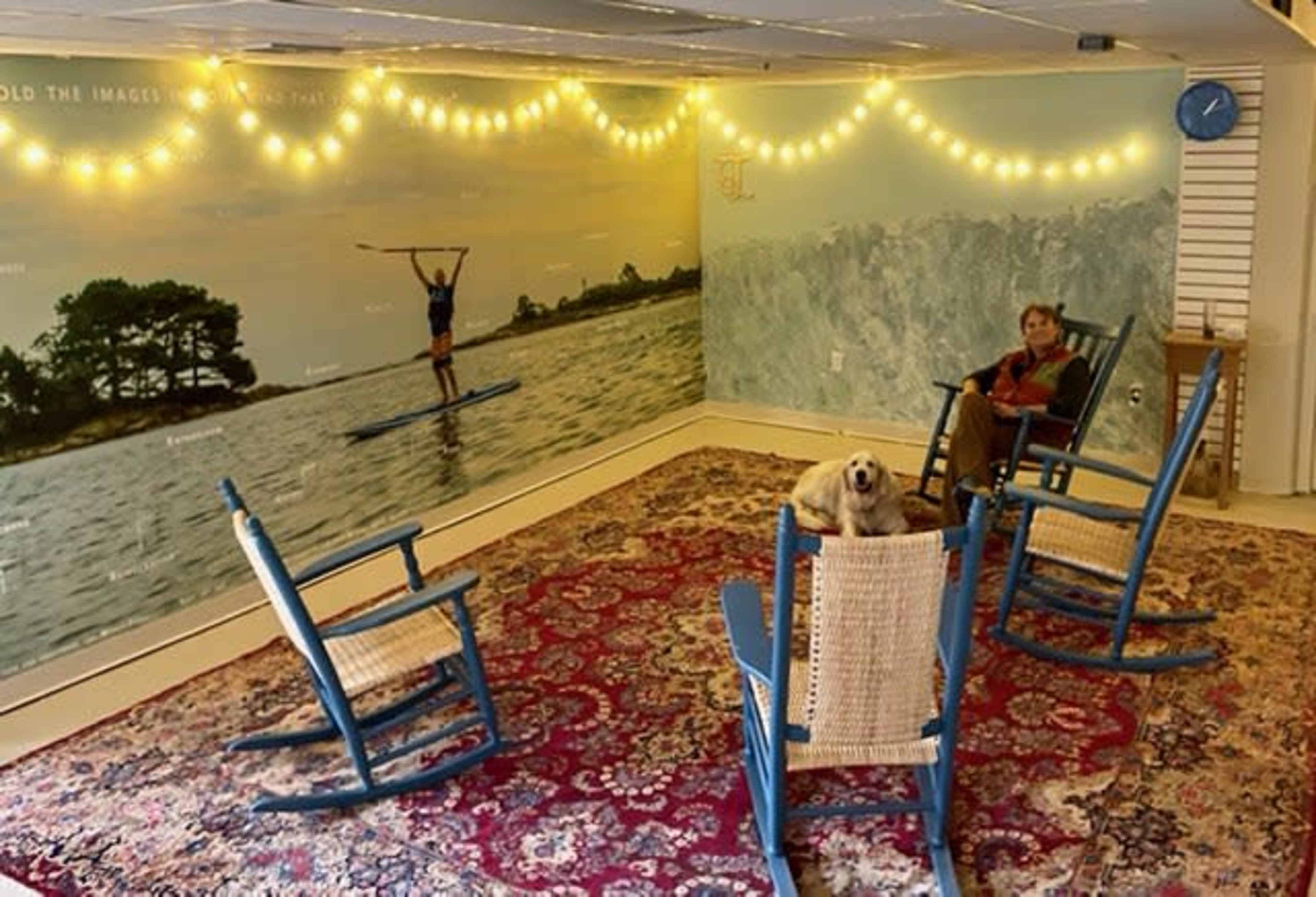 A room with four blue rocking chairs arranged around a patterned rug, a dog sitting on one of the chairs, and a mural depicting a person paddleboarding on a lake.