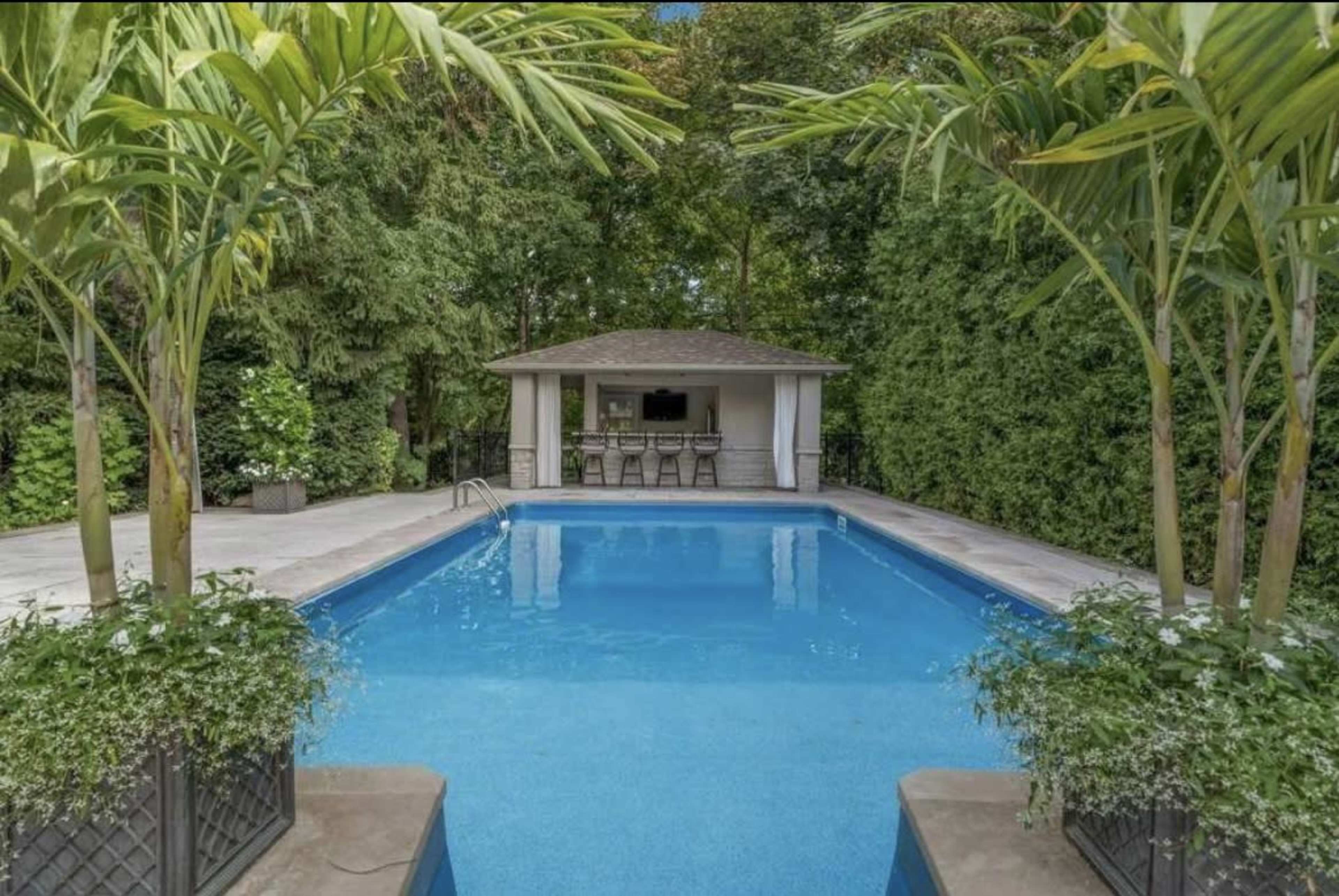 The image shows a clear blue swimming pool surrounded by lush greenery and a gazebo with seating in the background.