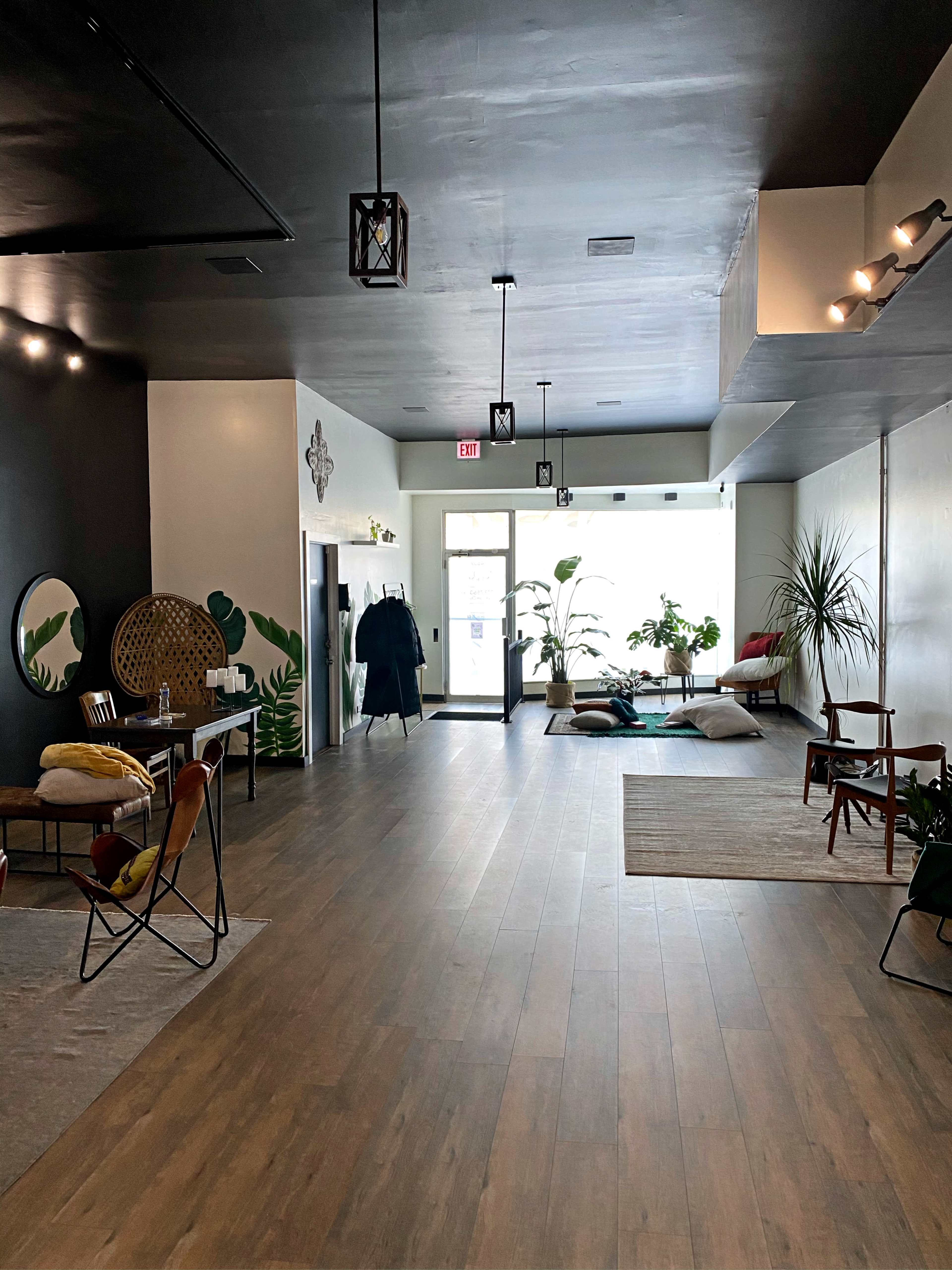 A spacious interior with wooden flooring, plants, various seating arrangements, and a large entrance filled with natural light.