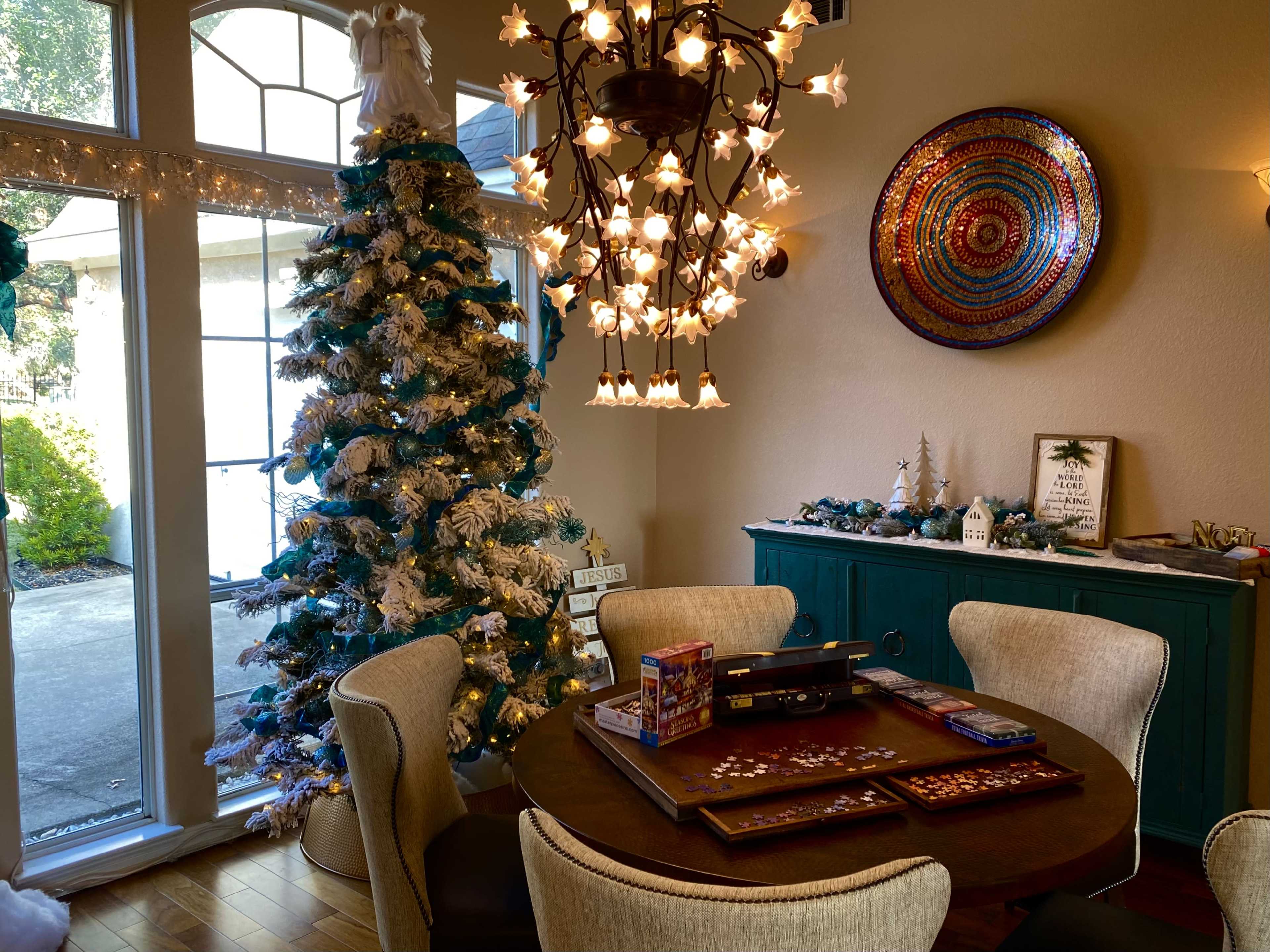 A decorated Christmas tree with white and blue ornaments stands next to a dining table surrounded by chairs, beneath a chandelier in a brightly lit room.