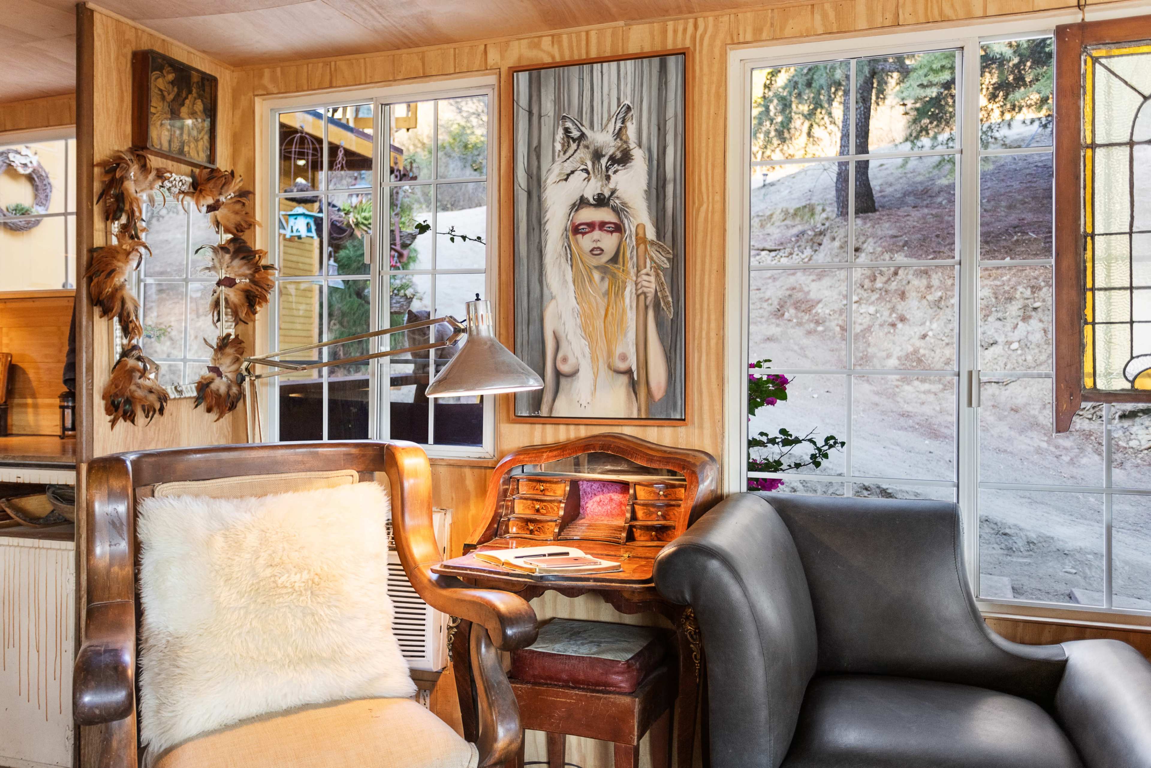 The interior of a cabin features a wooden chair with a fur cushion, a vintage writing desk, and large windows showcasing a view of the outdoors.