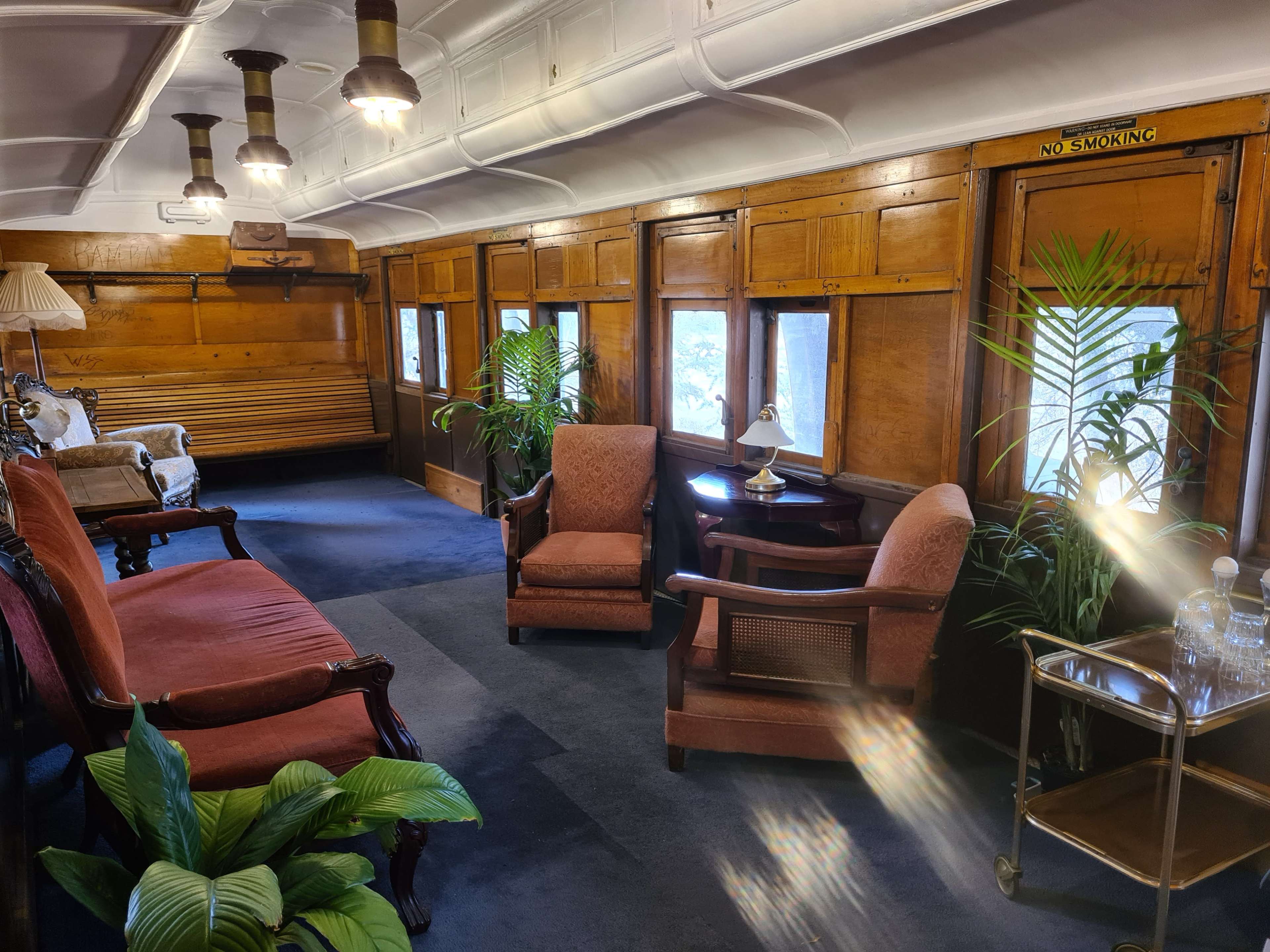 The interior of a vintage train car features wooden paneling, vintage seating, and plants near a small table with a lamp.