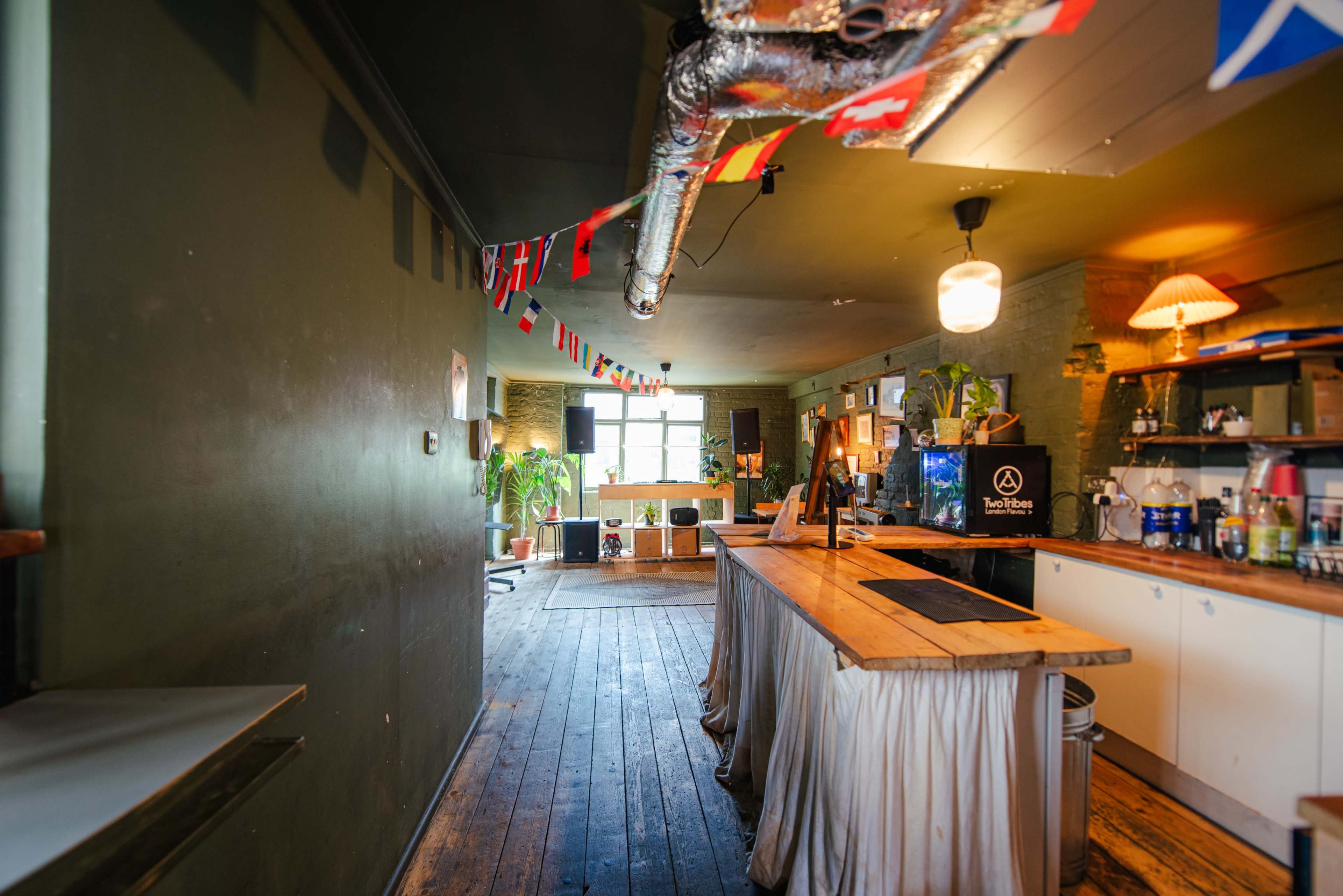 The image shows a wide shot of a cozy café interior with wooden flooring, a bar counter, and decorative flags hanging from the ceiling.