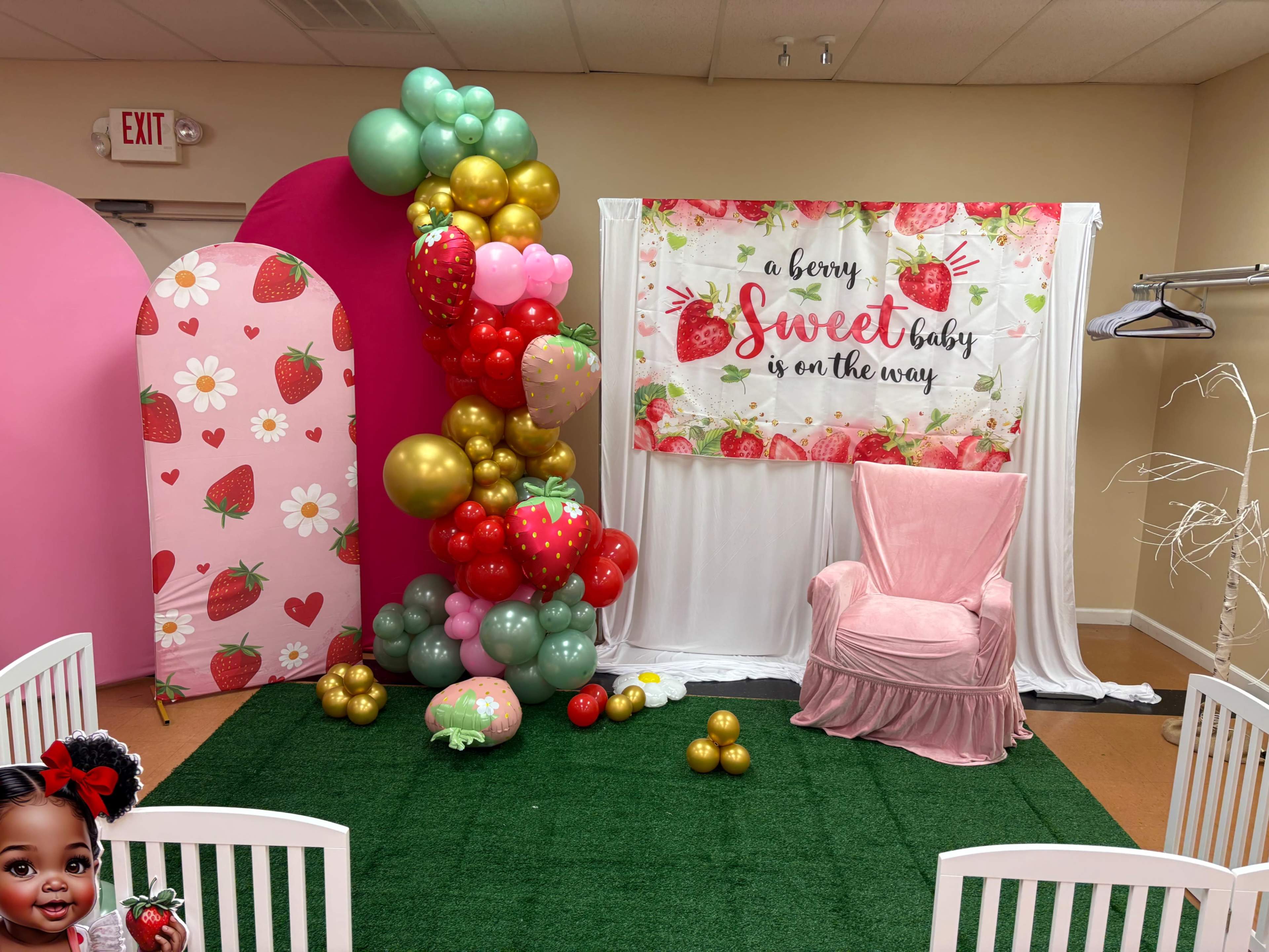 The image shows a decorated event space featuring a grassy area, a pink chair, vibrant balloon arrangements, and a backdrop with a strawberry-themed banner.