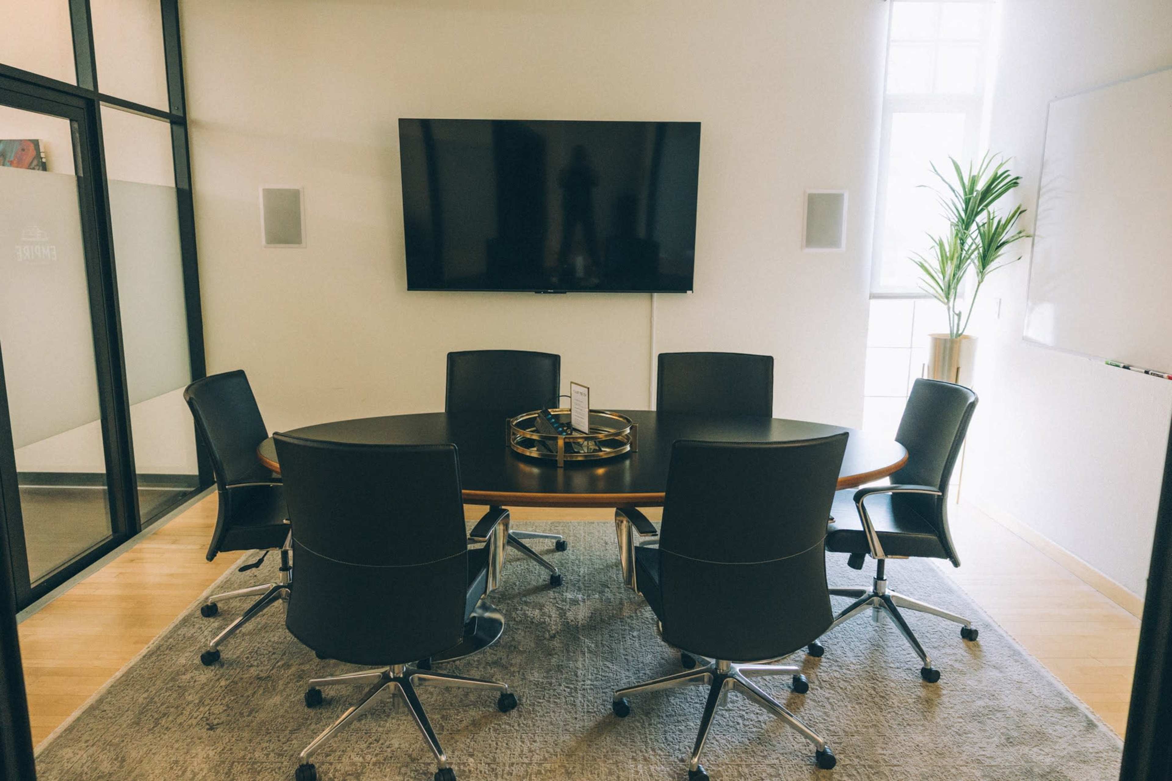 A conference room features a round table surrounded by five black chairs, with a television mounted on the wall and a plant in the corner.