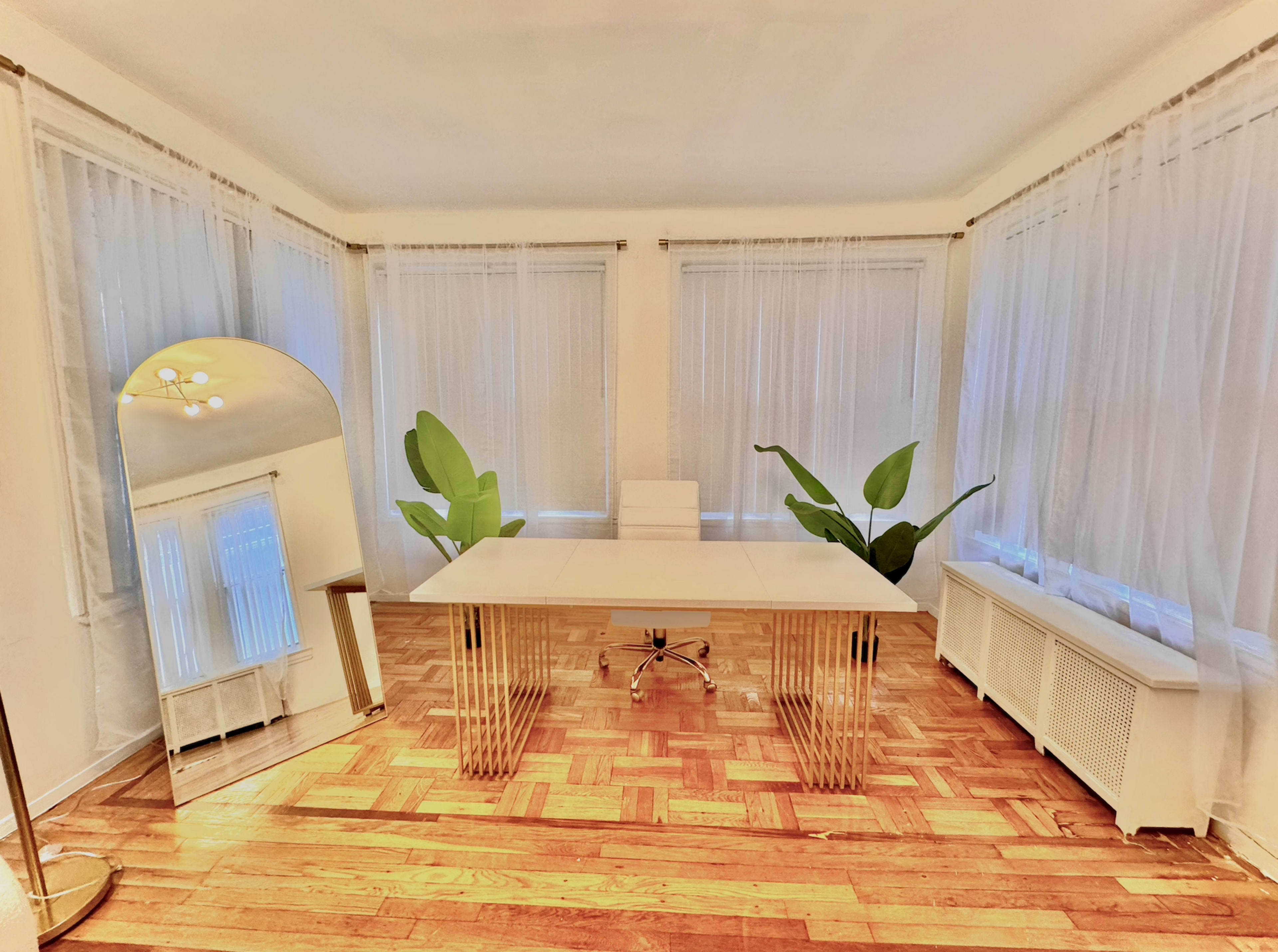 A minimalistic office space with a white desk, a white chair, large windows covered by sheer curtains, and two potted plants.