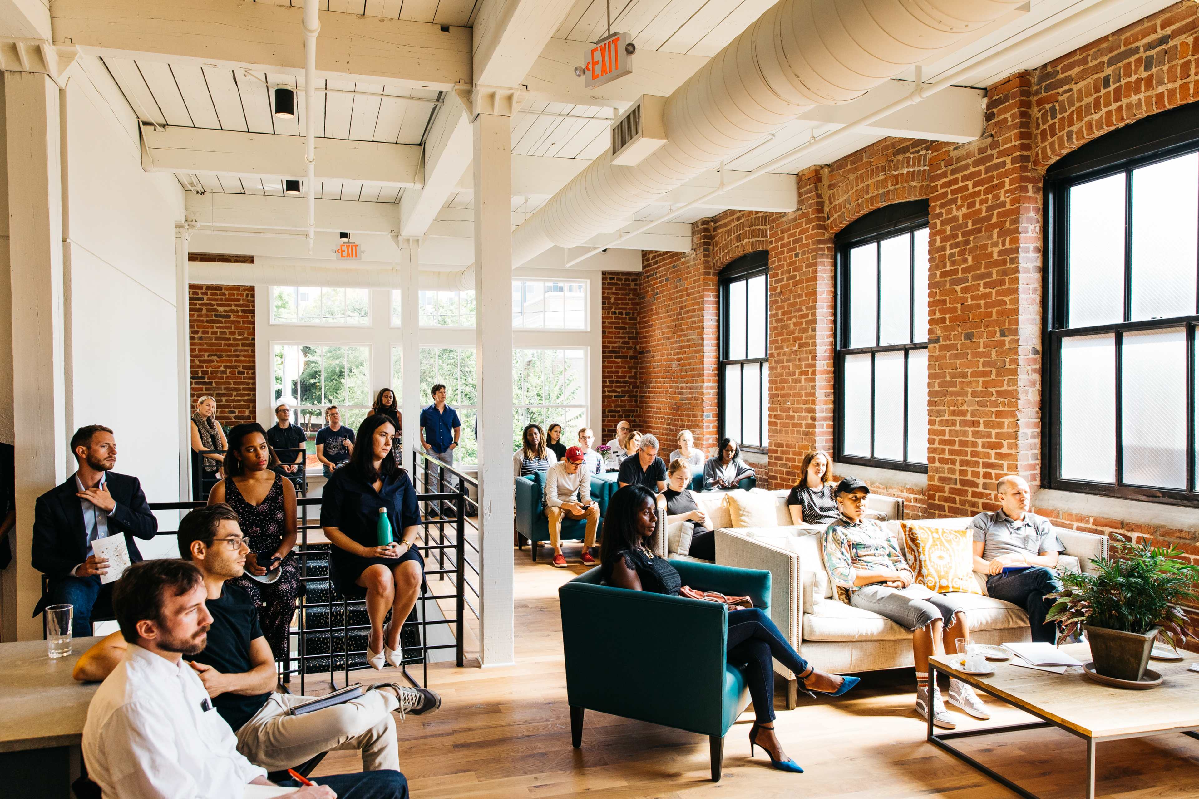 A diverse group of people is seated in a modern, brick-walled room, engaged in a presentation or discussion.