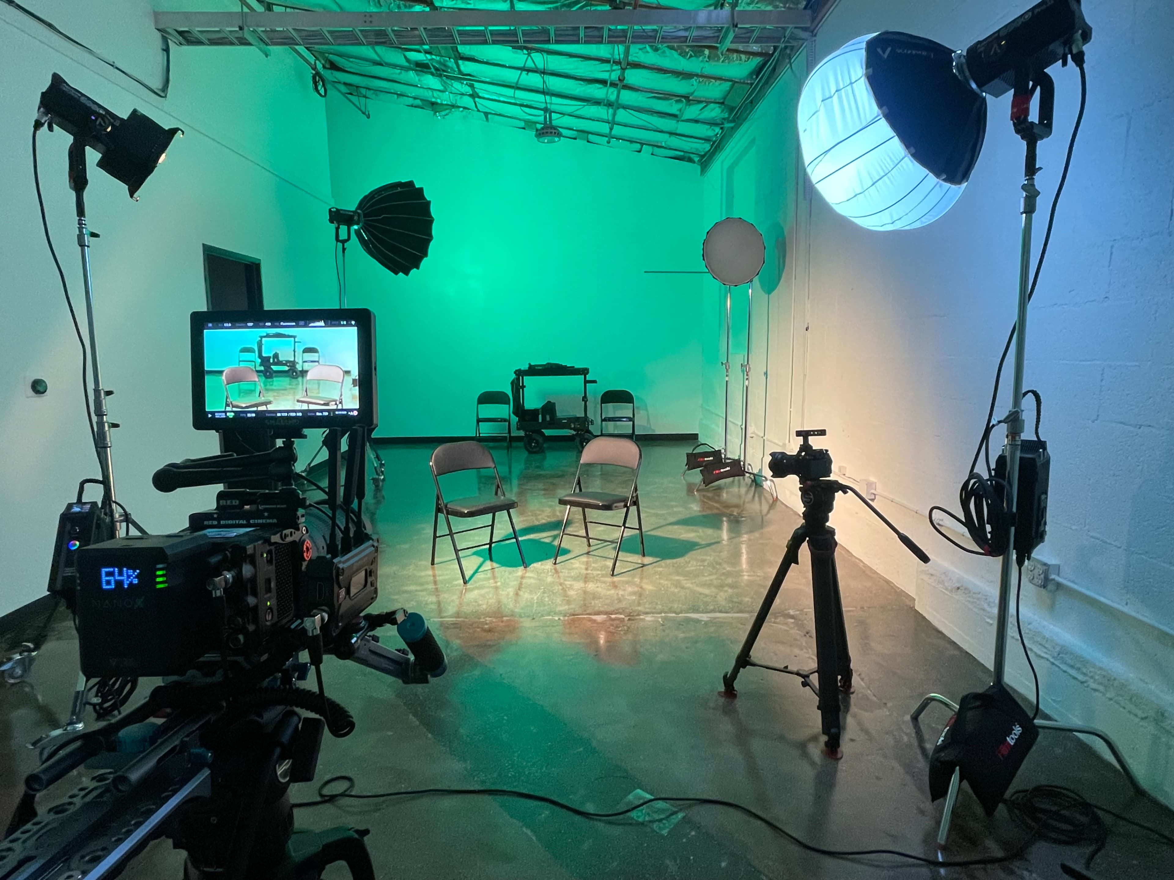 The image shows a photography or video production setup in a studio with two empty chairs, various studio lights, and cameras positioned on tripods.