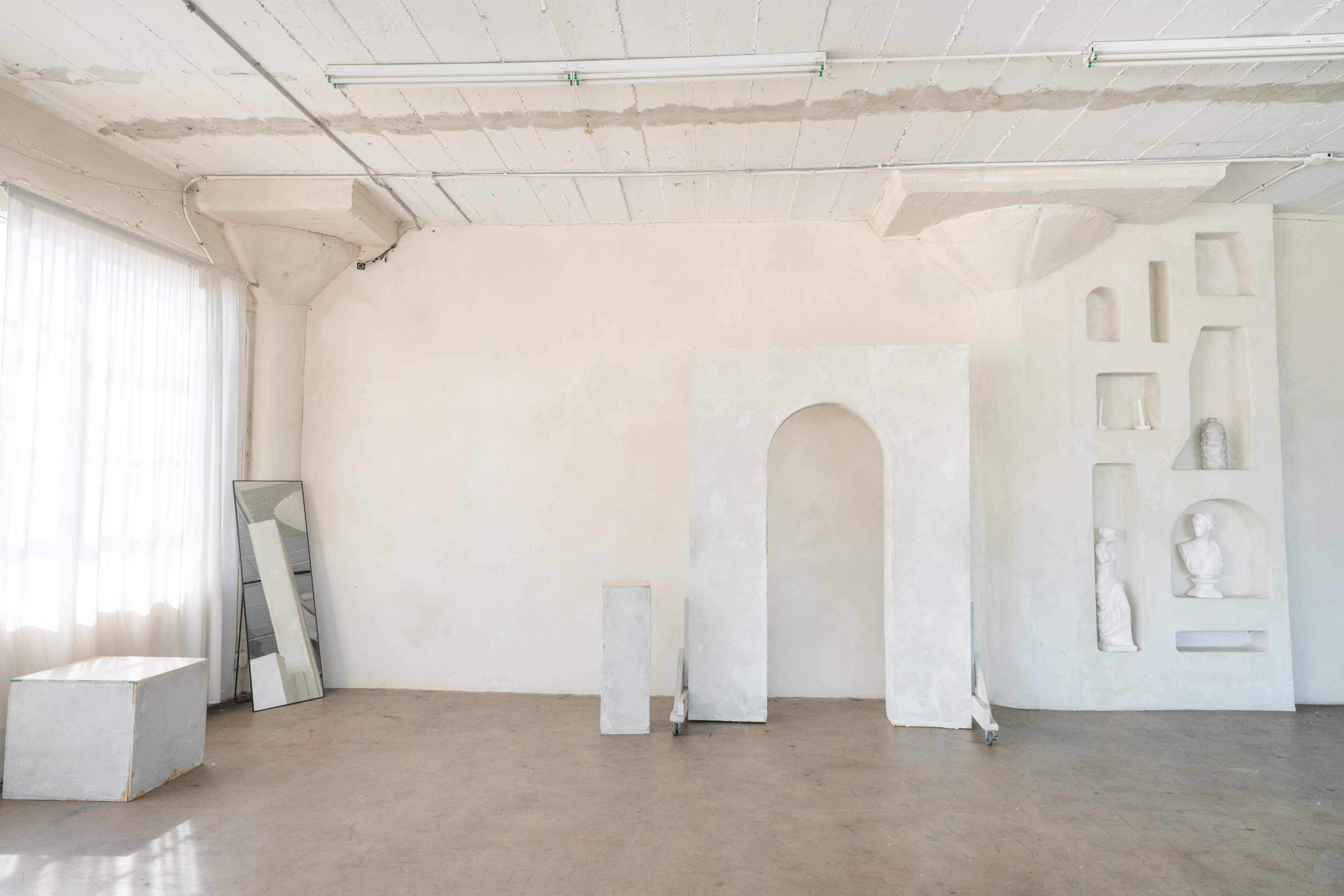 The image shows a minimalist interior space with white walls, a large window with sheer curtains, and several sculptural elements, including a tall arch and a pedestal, alongside a mirror and a grey cube.