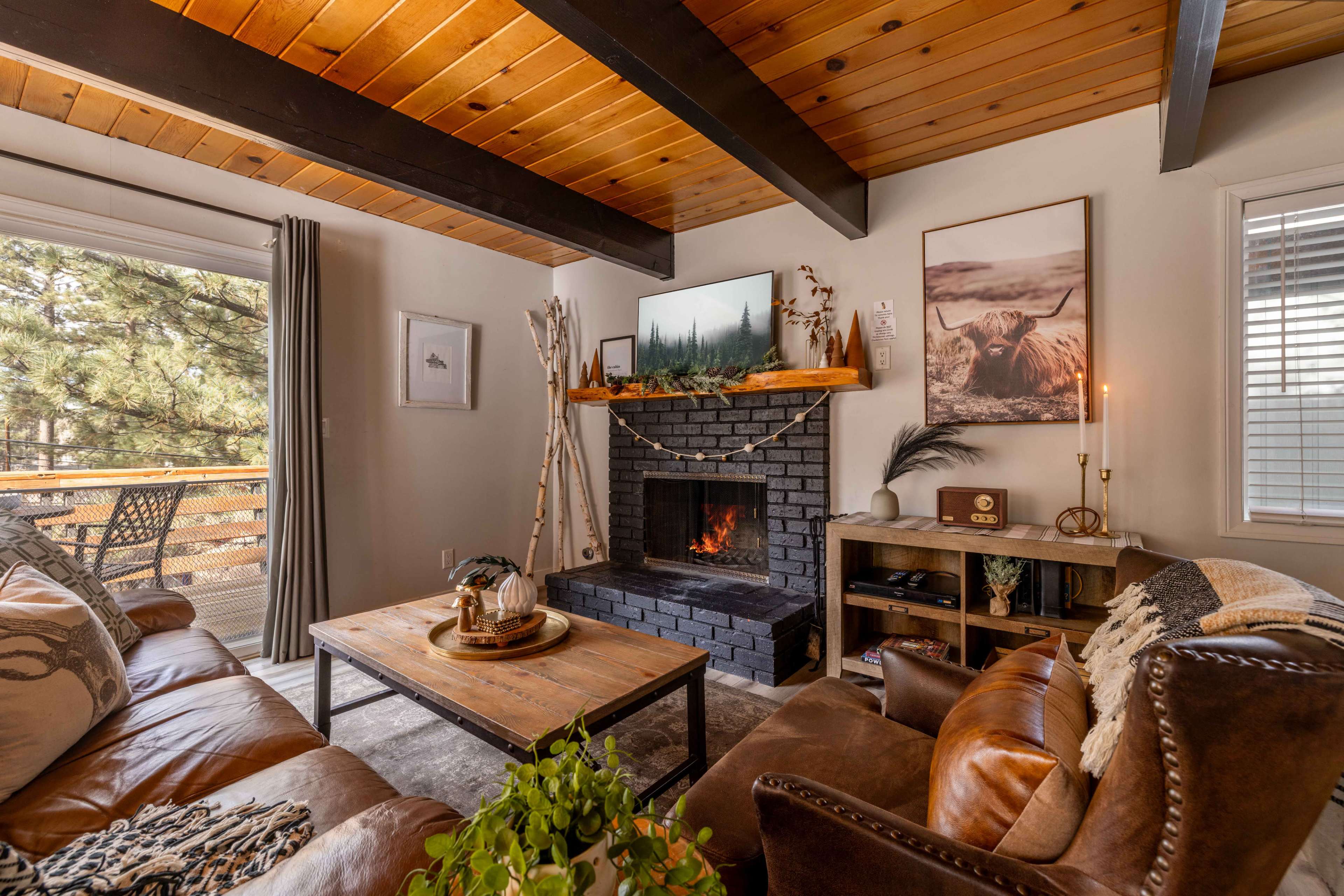 A cozy living room features a dark brick fireplace with a crackling fire, surrounded by wood-beam ceilings and natural light from large windows.