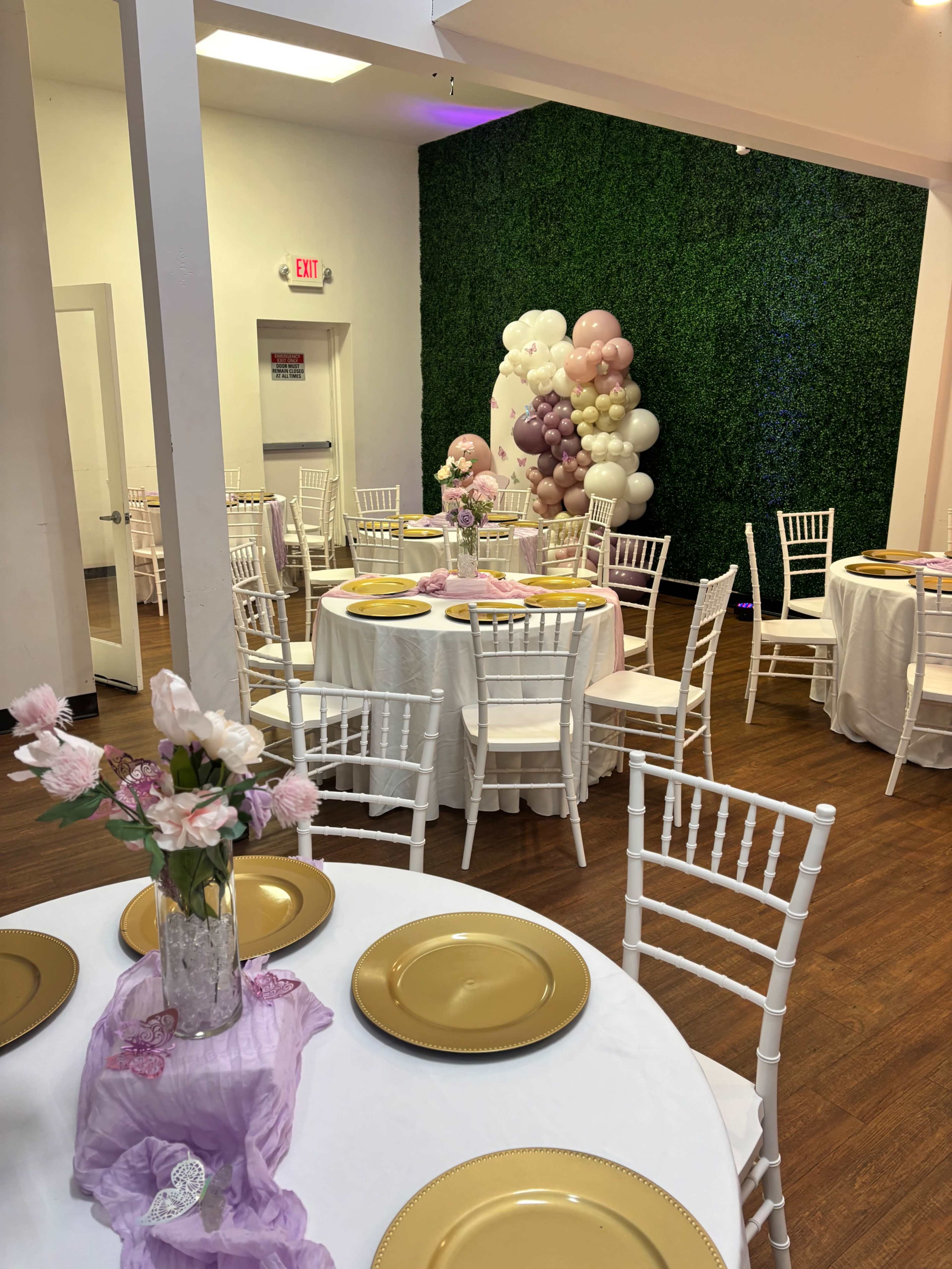 The image shows a decorated event space with circular tables set with white tablecloths, gold plates, and floral centerpieces, along with a vibrant balloon display against a green wall.