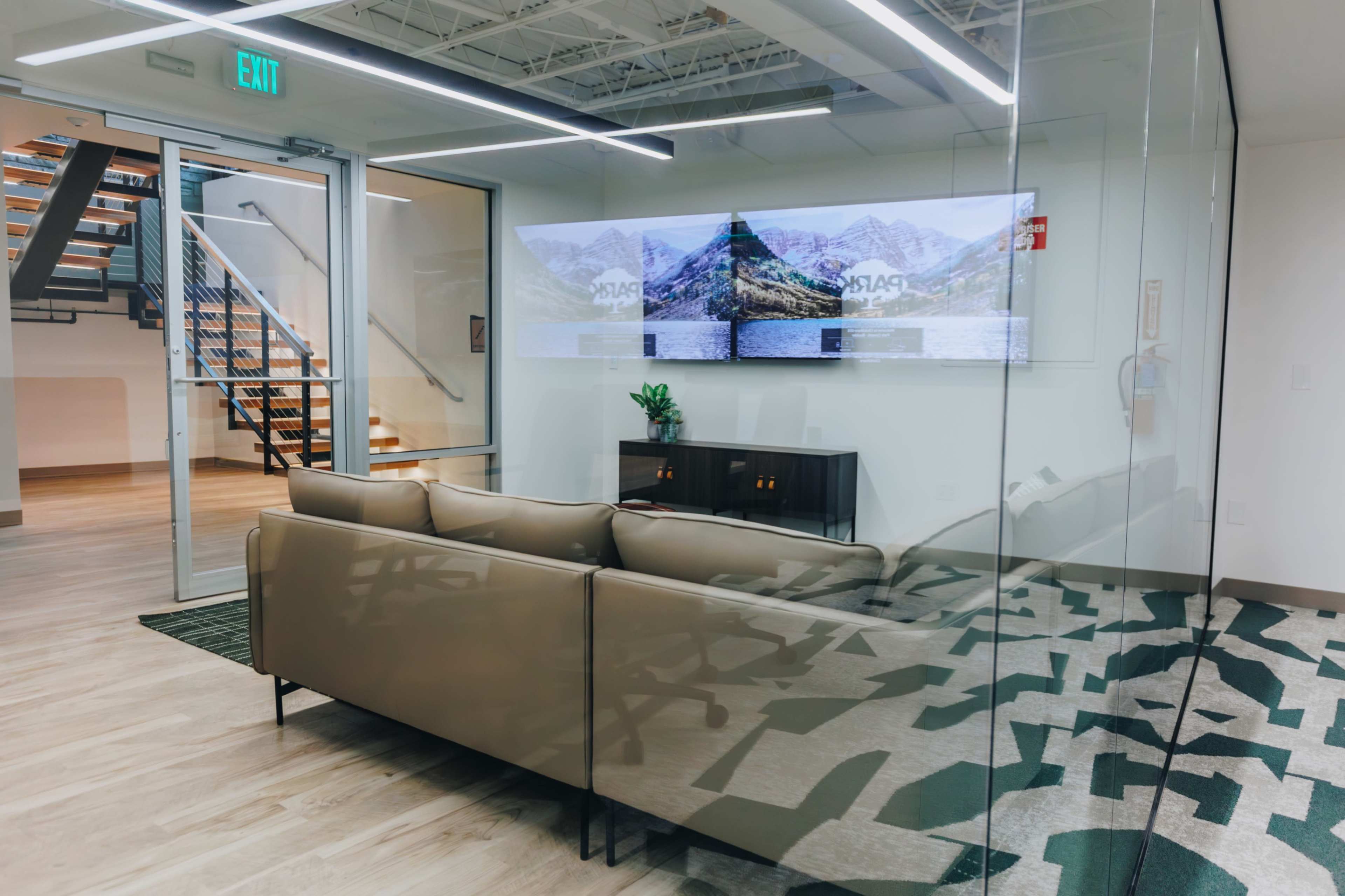 The image shows a modern office lounge area with a glass wall, a sofa, and a large screen displaying a scenic mountain view.