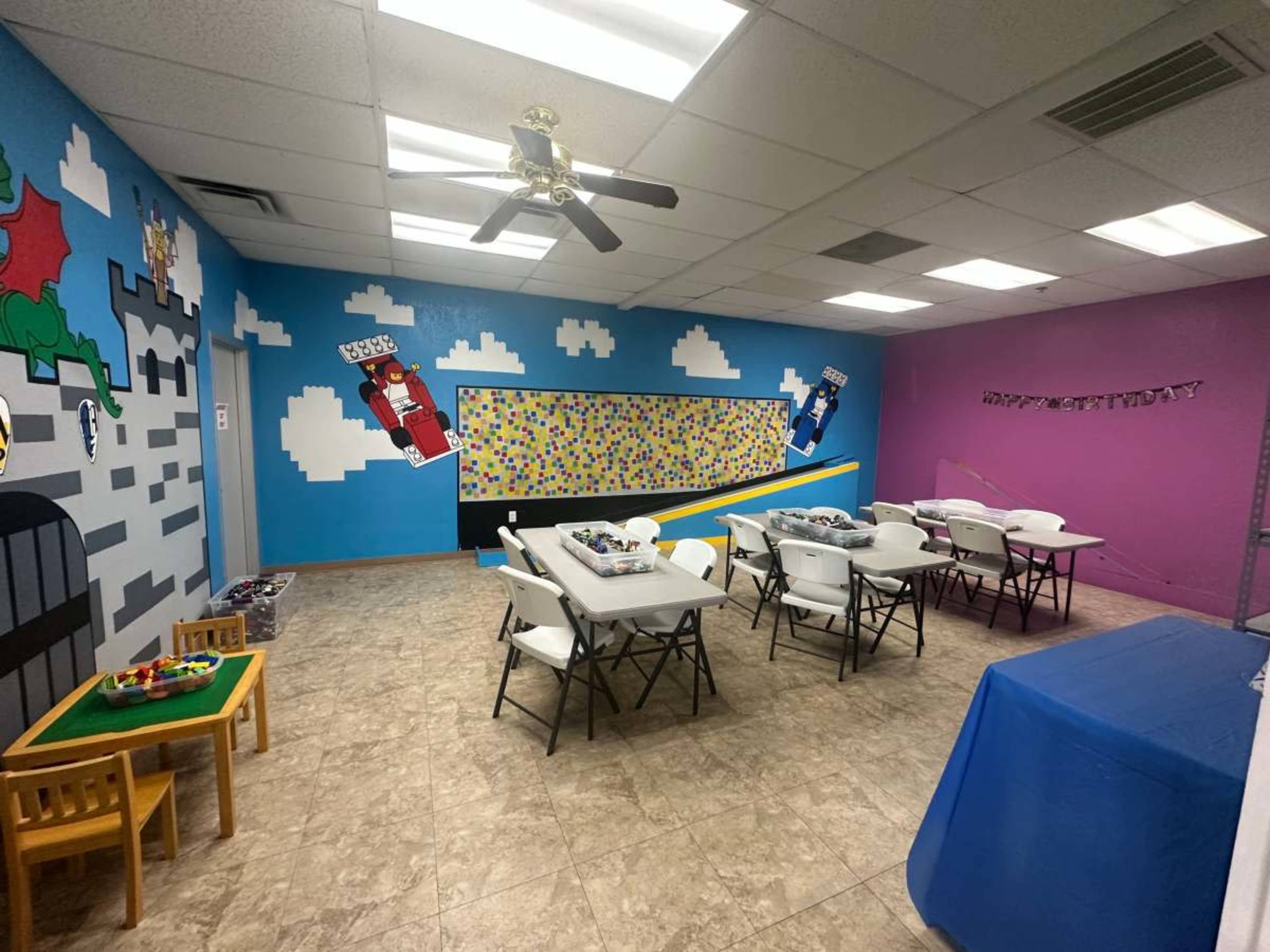 The image shows a colorful activity room with tables and chairs arranged for a gathering, featuring murals of superheroes and a bulletin board.