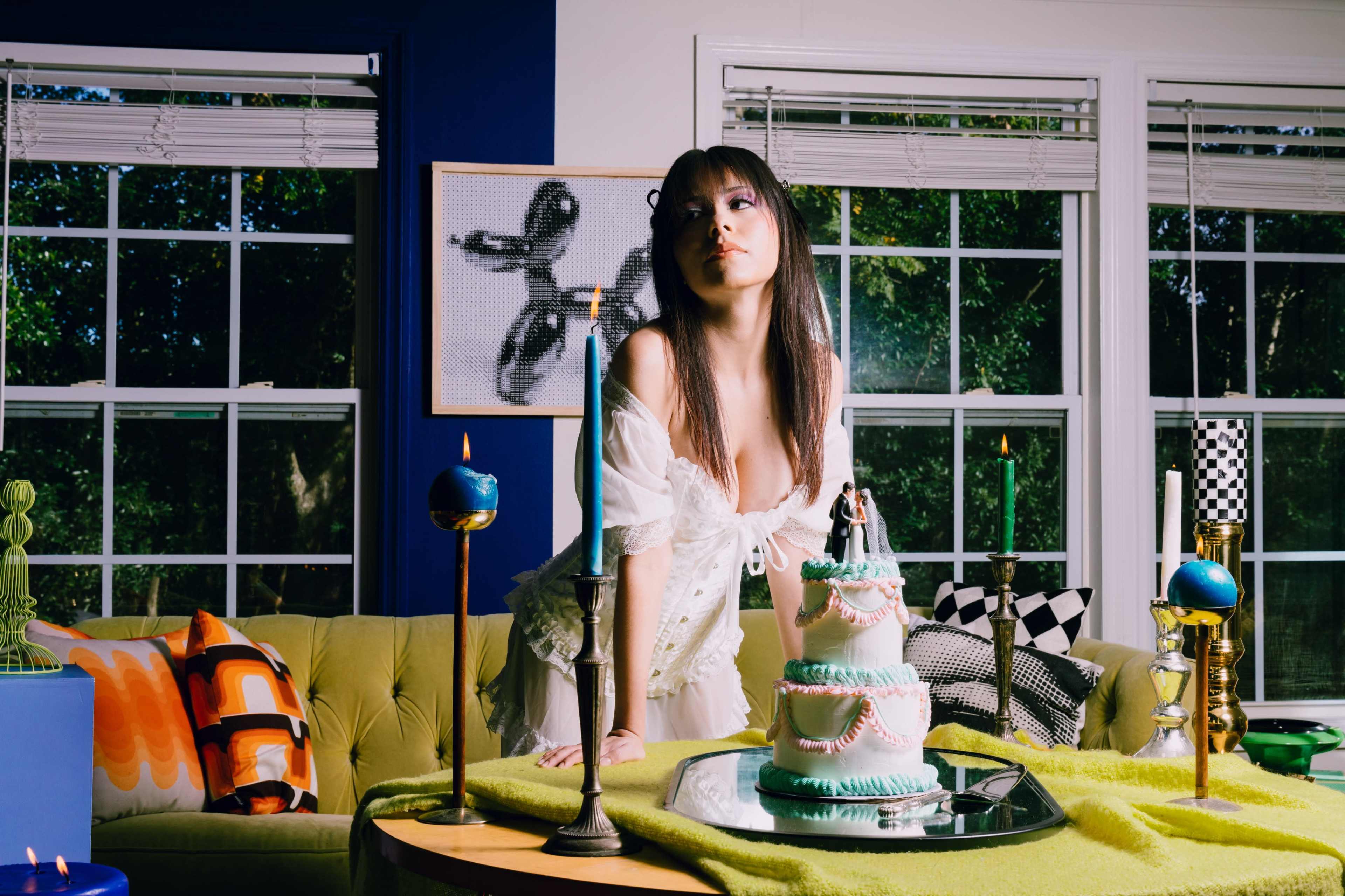 A person stands beside a three-tiered cake on a table adorned with candles and colorful decor in a well-lit room with large windows.