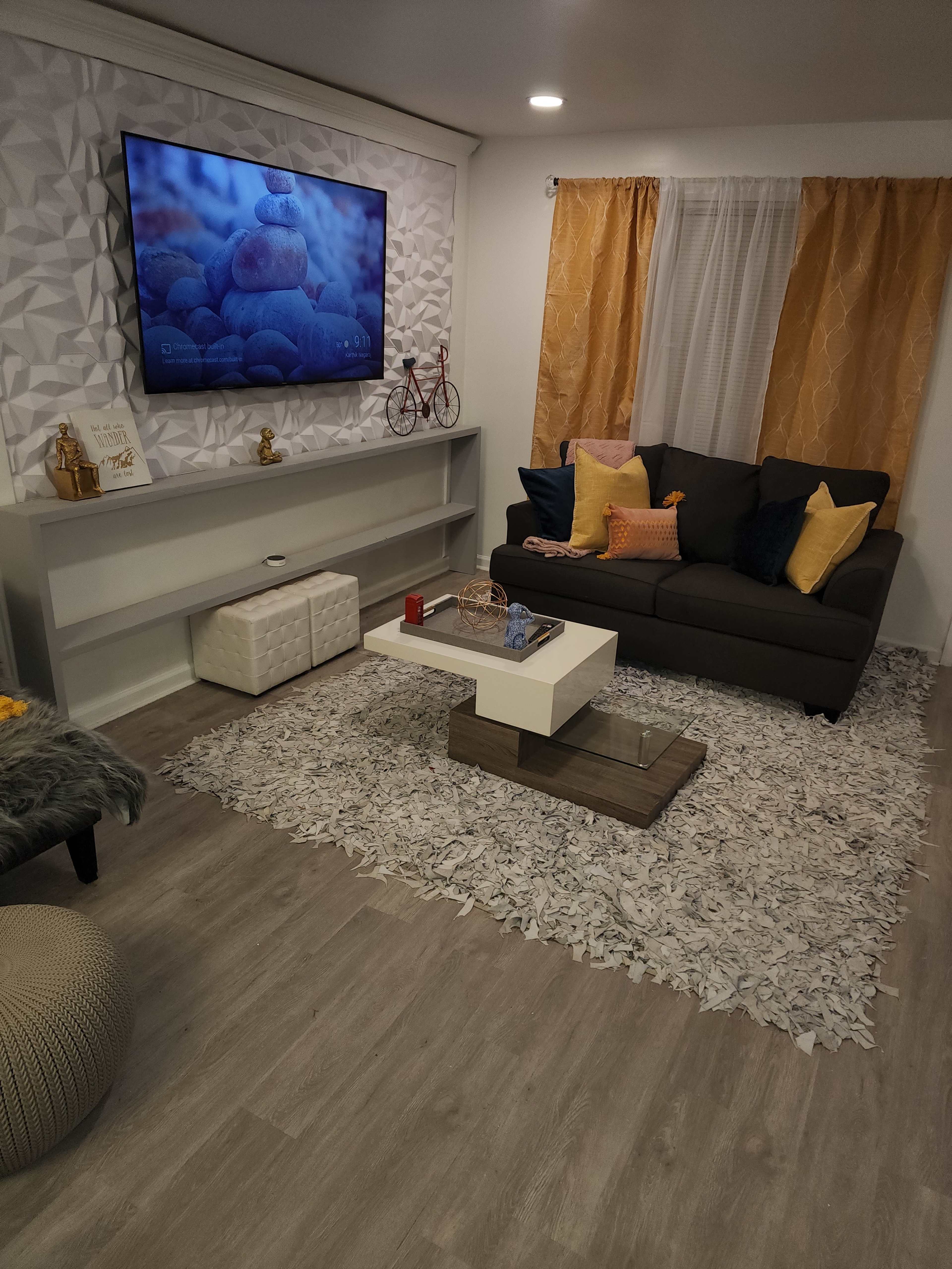 The room features a cozy seating area with a dark sofa, a coffee table, a TV mounted on the wall, and decorative elements like cushions and a rug.