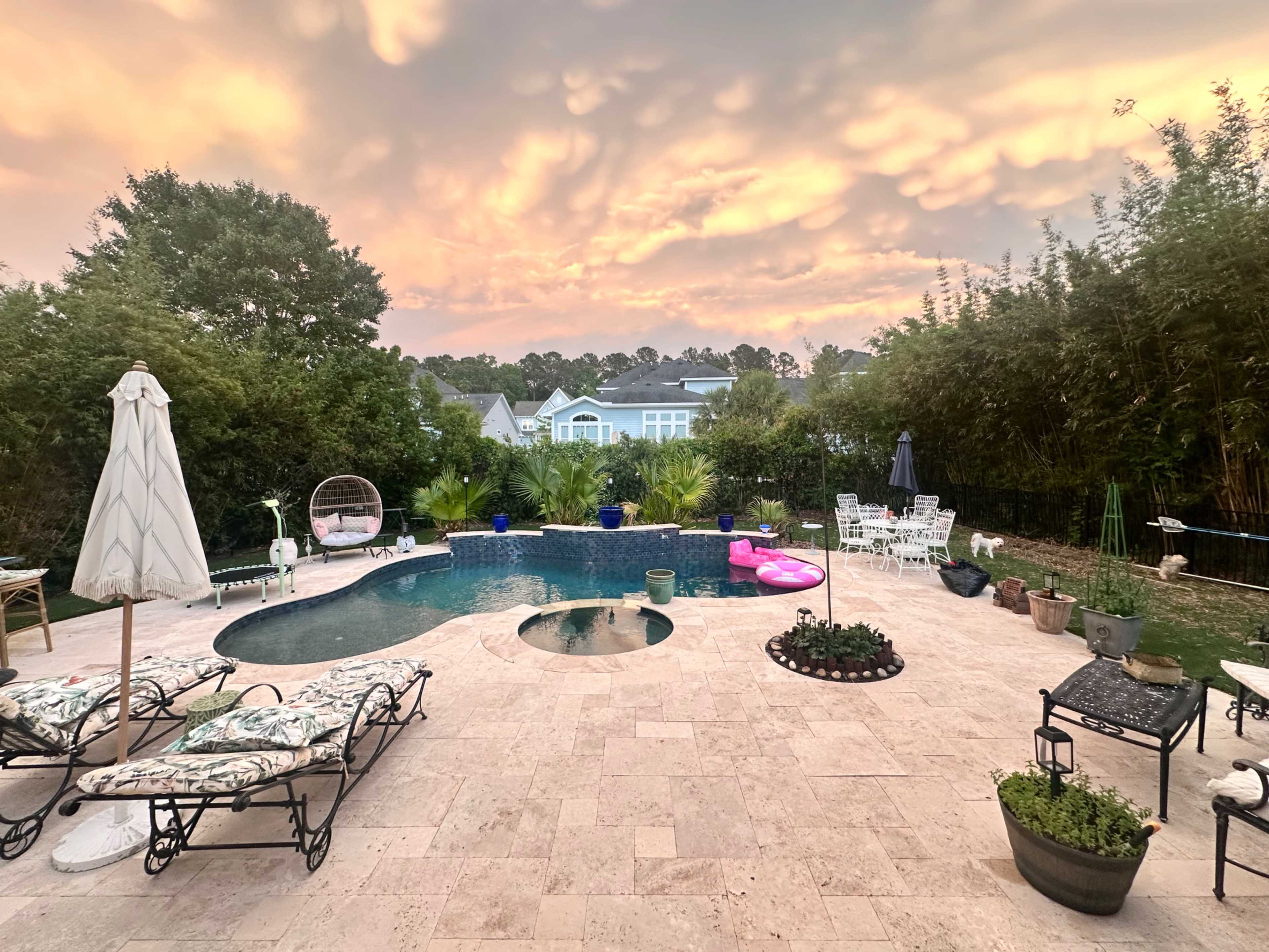 The image shows a pool area surrounded by patio furniture and lush greenery under a colorful sky at sunset.