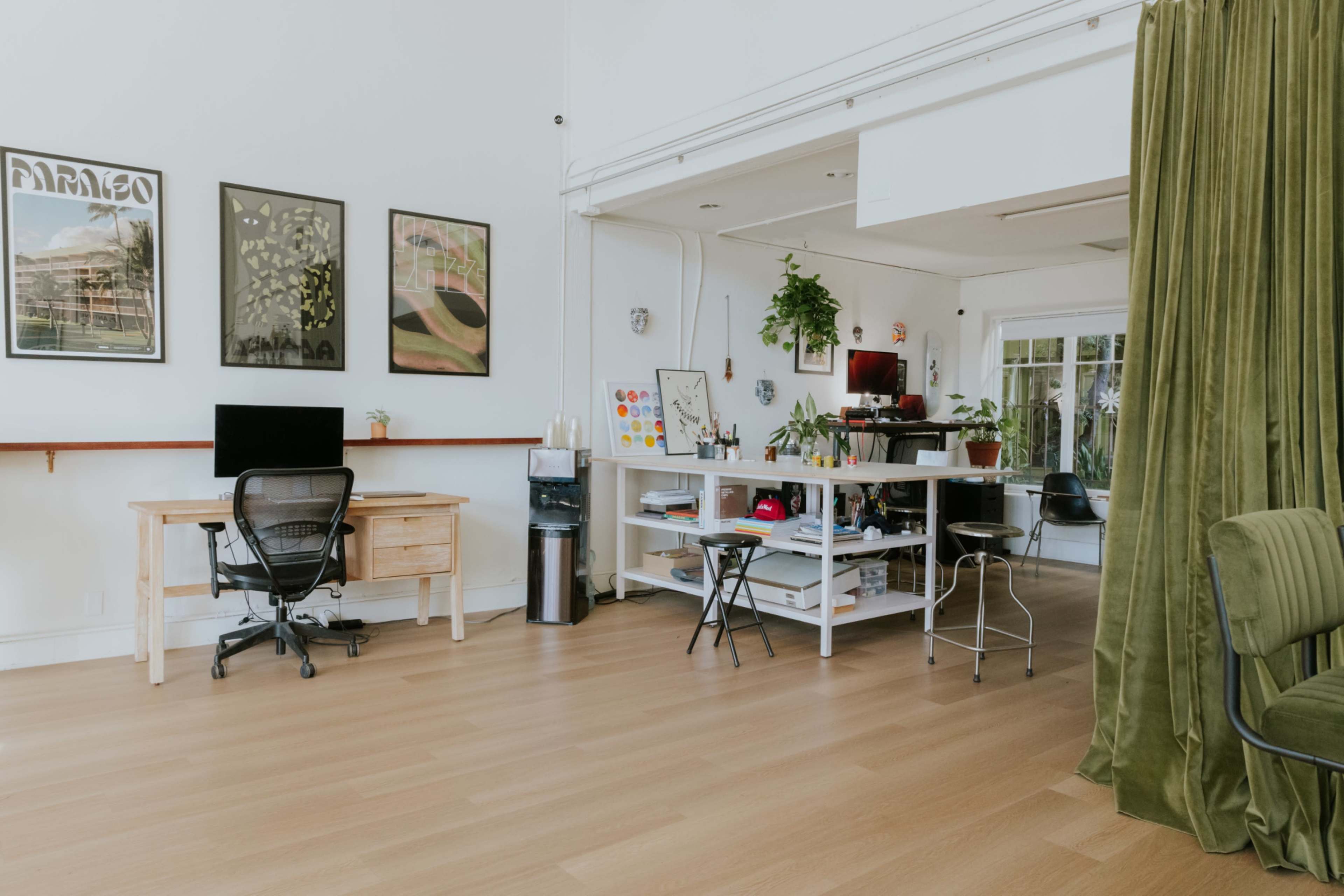 A bright office space features a wooden desk with a chair, a computer monitor, and various plants, alongside shelves stacked with items and a curtain separating areas.