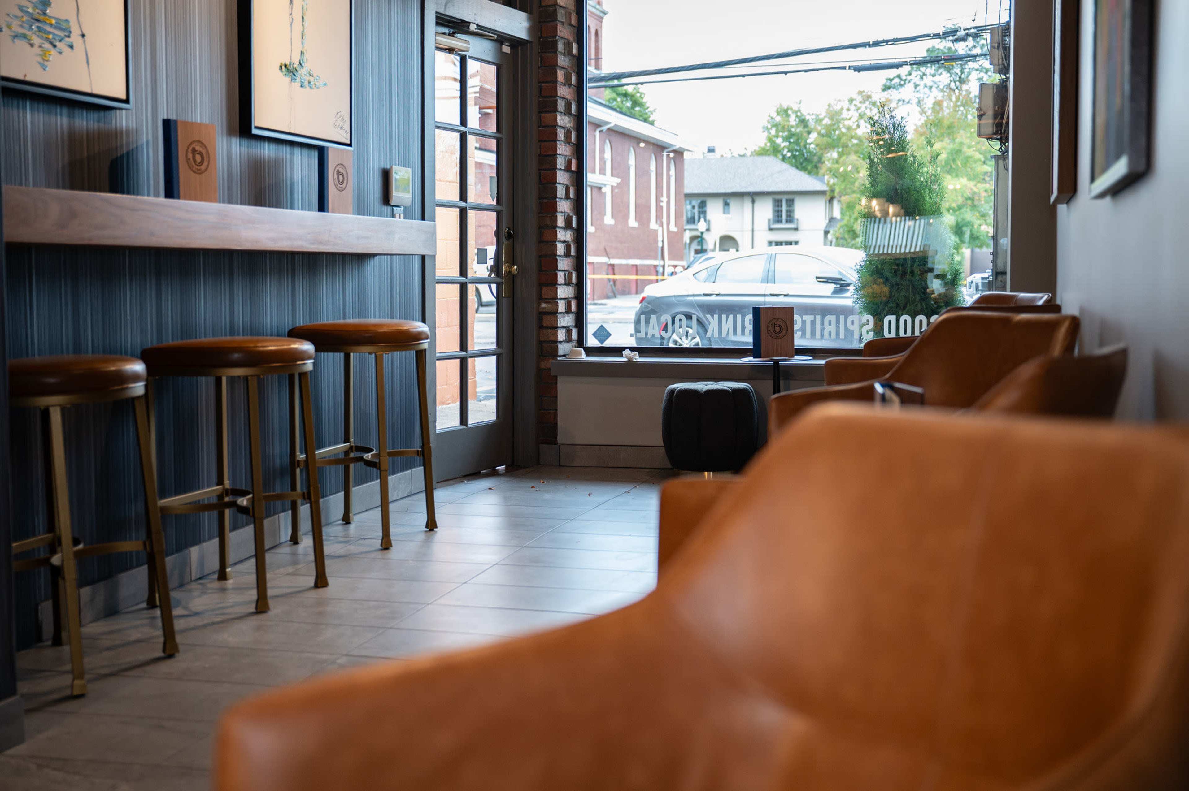 The image shows a modern interior of a coffee shop with wooden bar stools, brown leather chairs, and a glass storefront revealing a street with parked vehicles.