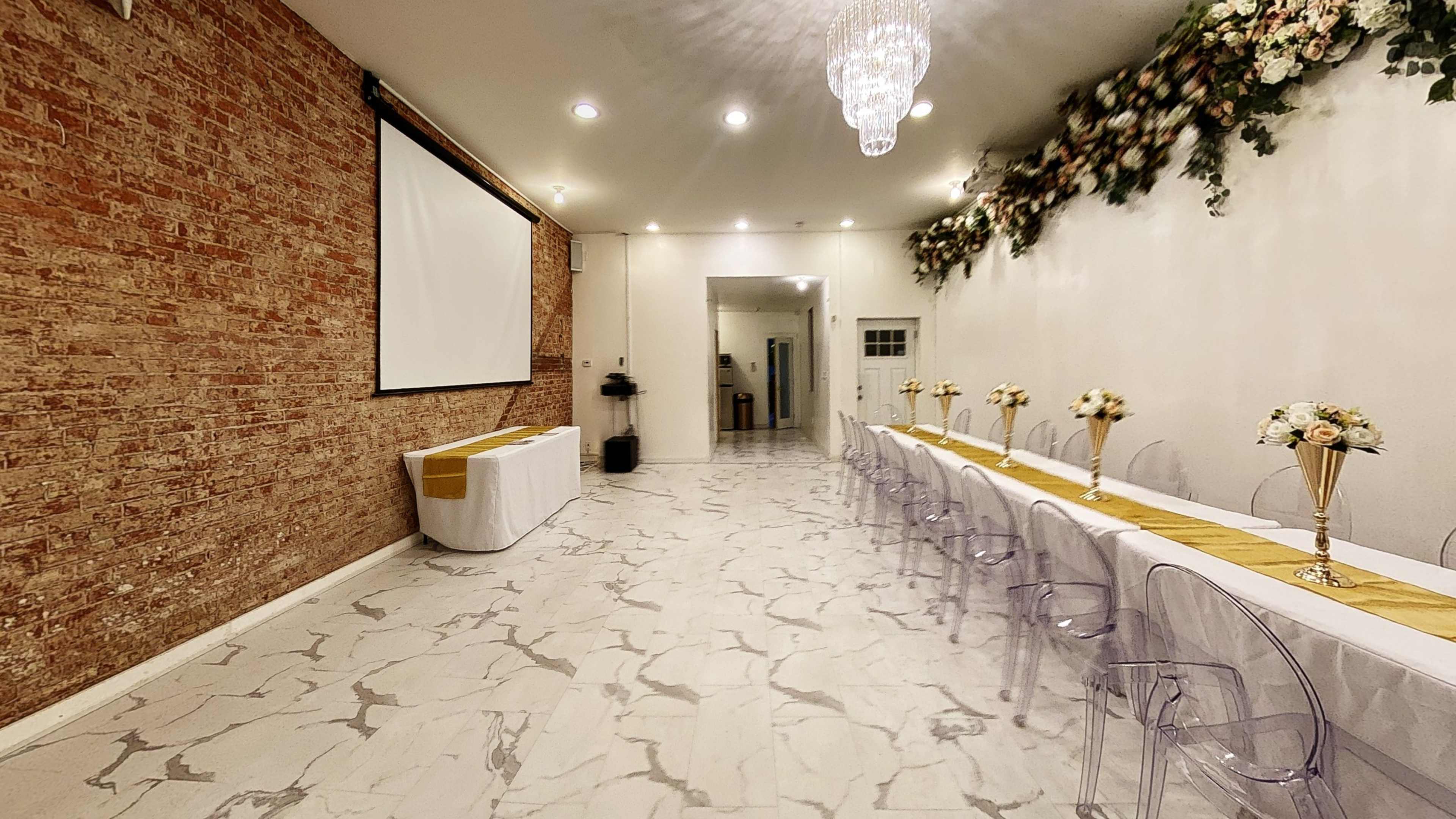 A spacious event room featuring a long table set with floral centerpieces, transparent chairs, a brick wall, and bright lighting.