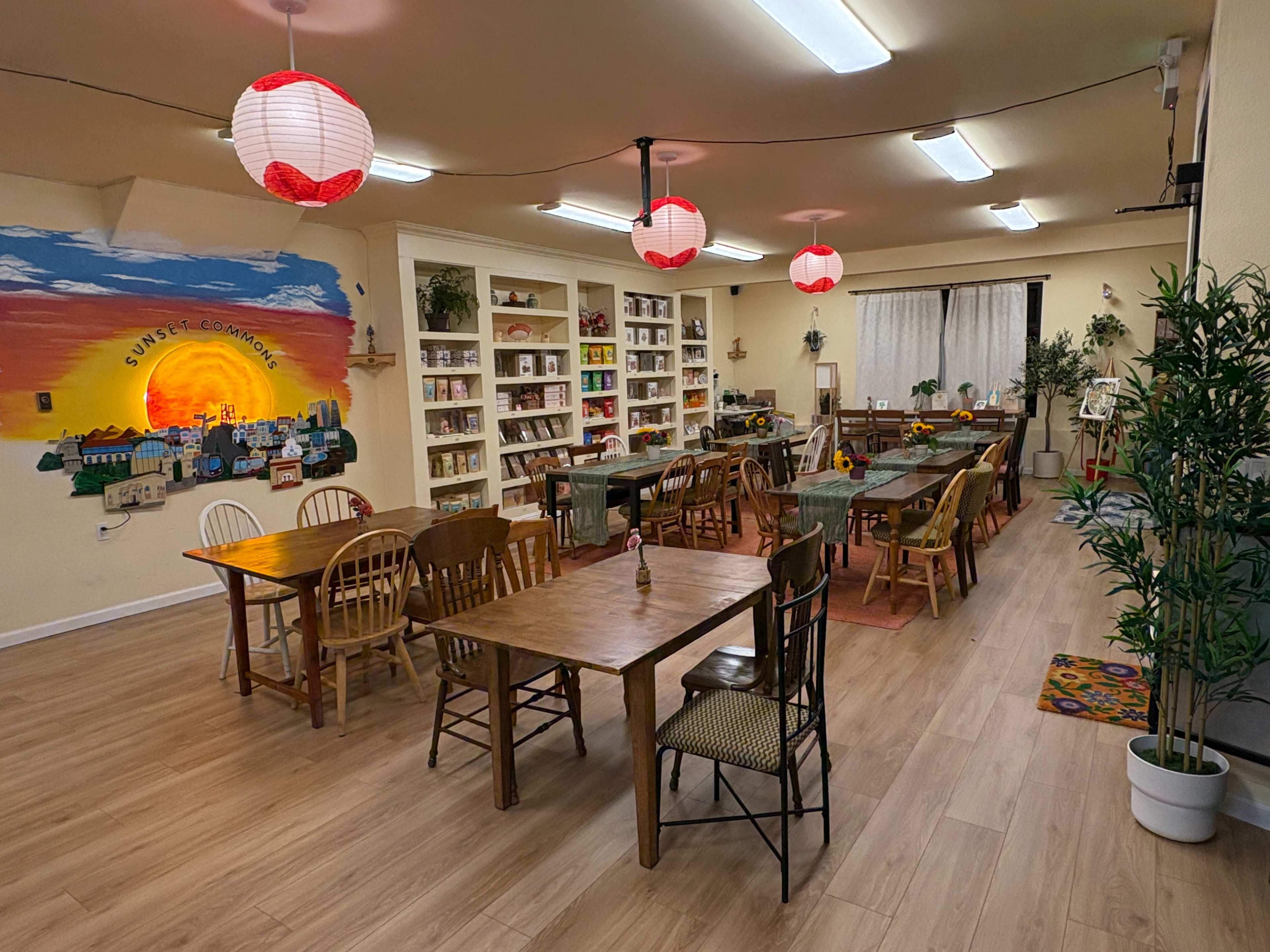 The image shows a spacious room filled with wooden tables and chairs, lined with shelves of books, and adorned with paper lanterns hanging from the ceiling.