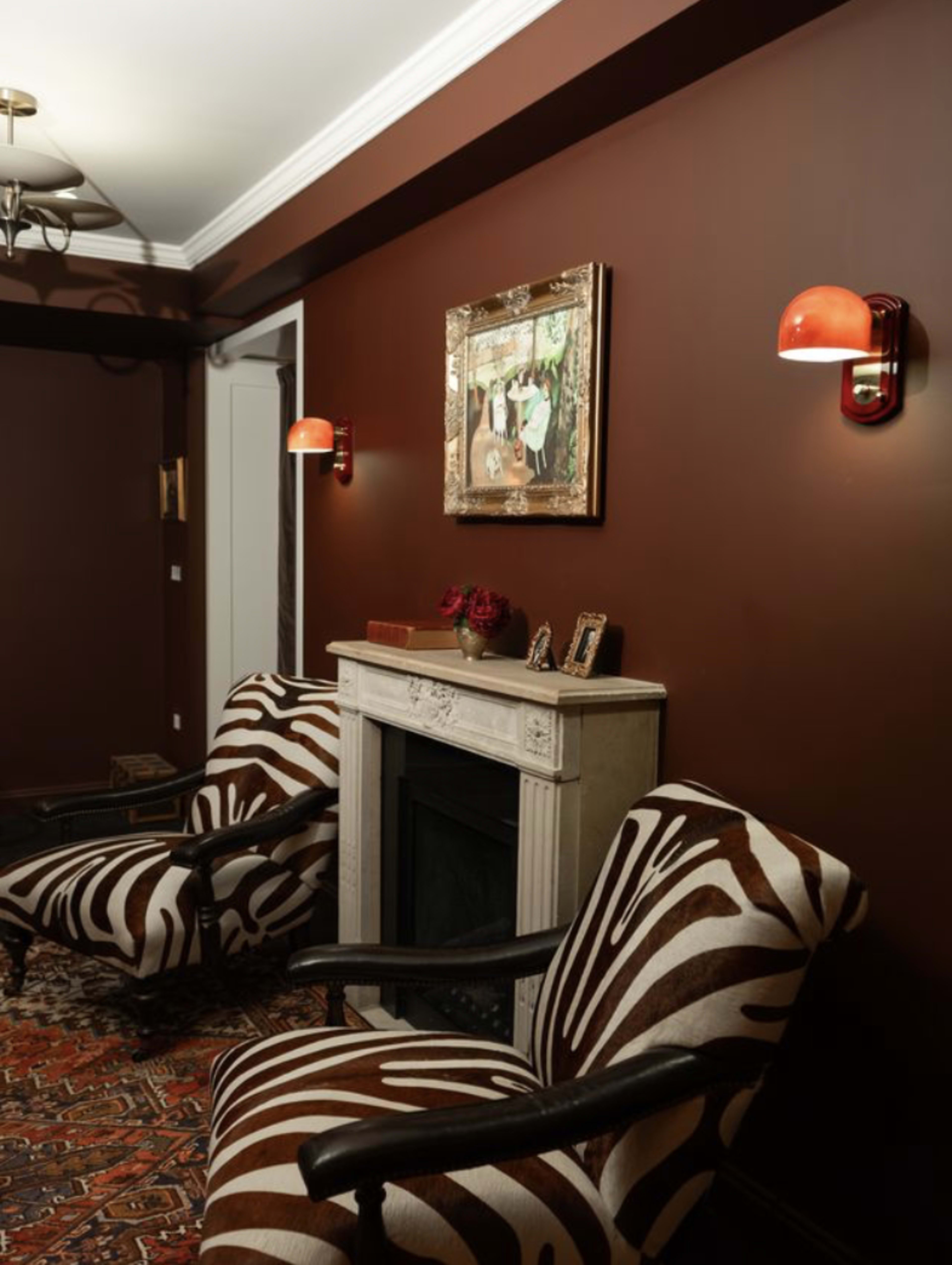 The image shows a room with dark brown walls, featuring two zebra-striped chairs flanking a white mantelpiece with a decorative painting above it.